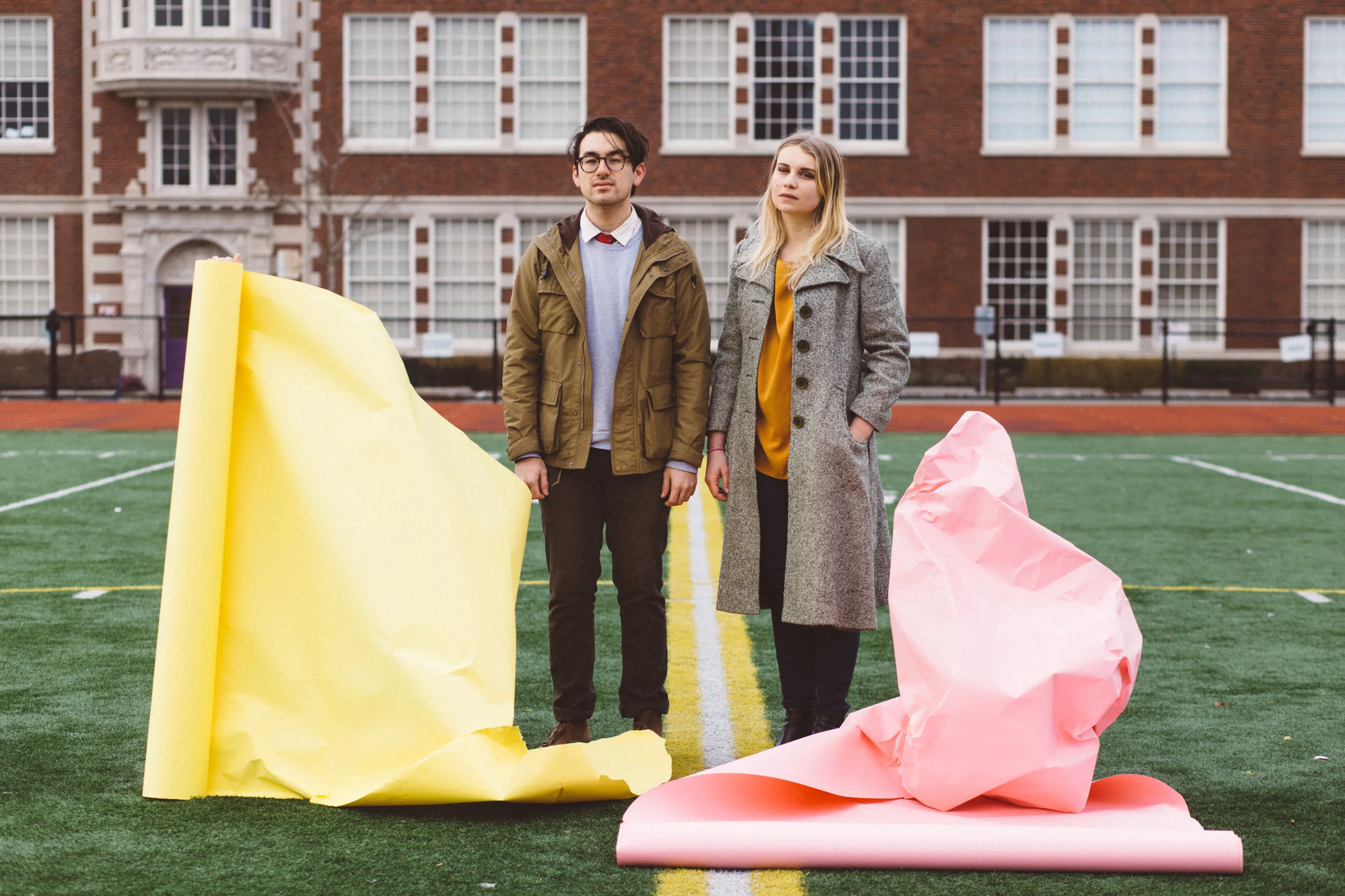 Two young adults standing on a sports field in front of a brick building, with large crumpled paper sculptures in yellow and pink on the ground nearby.