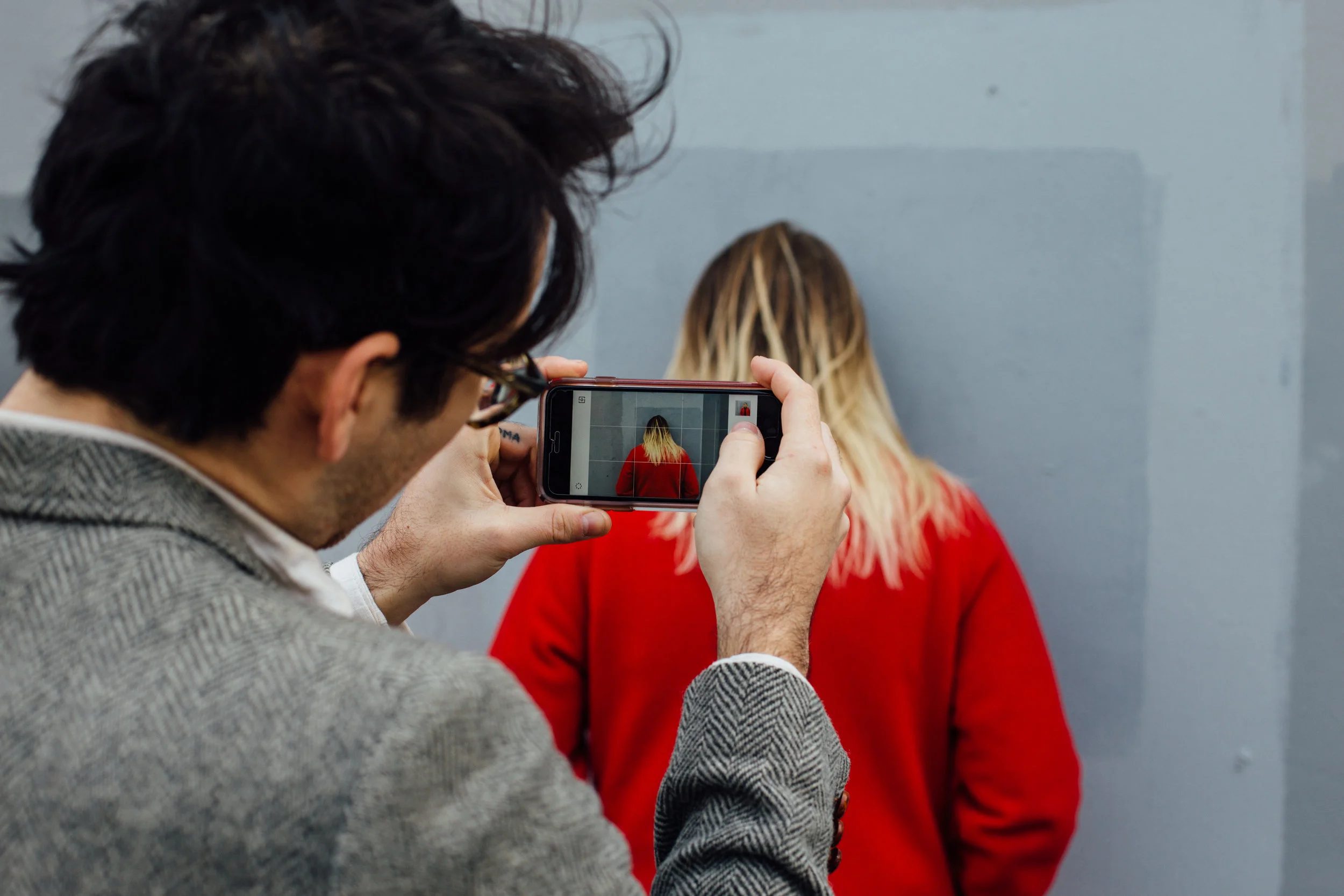 Man taking a photo of woman with yellow hair and red shirt against a gray wall.