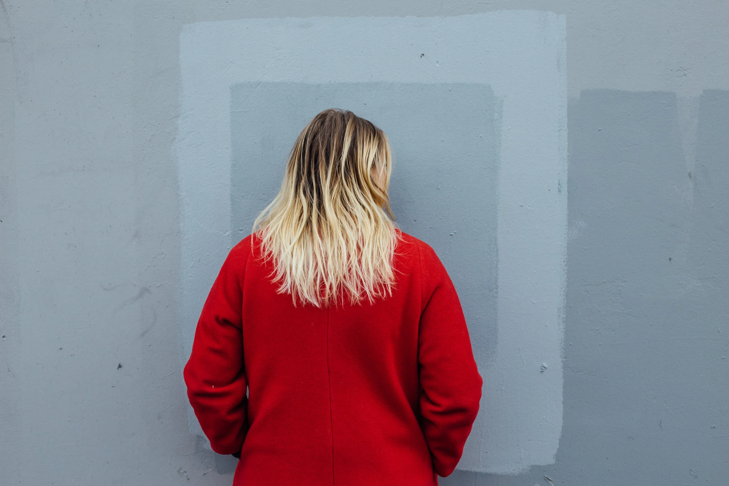 Person with blonde hair wearing a red coat facing a gray painted wall.