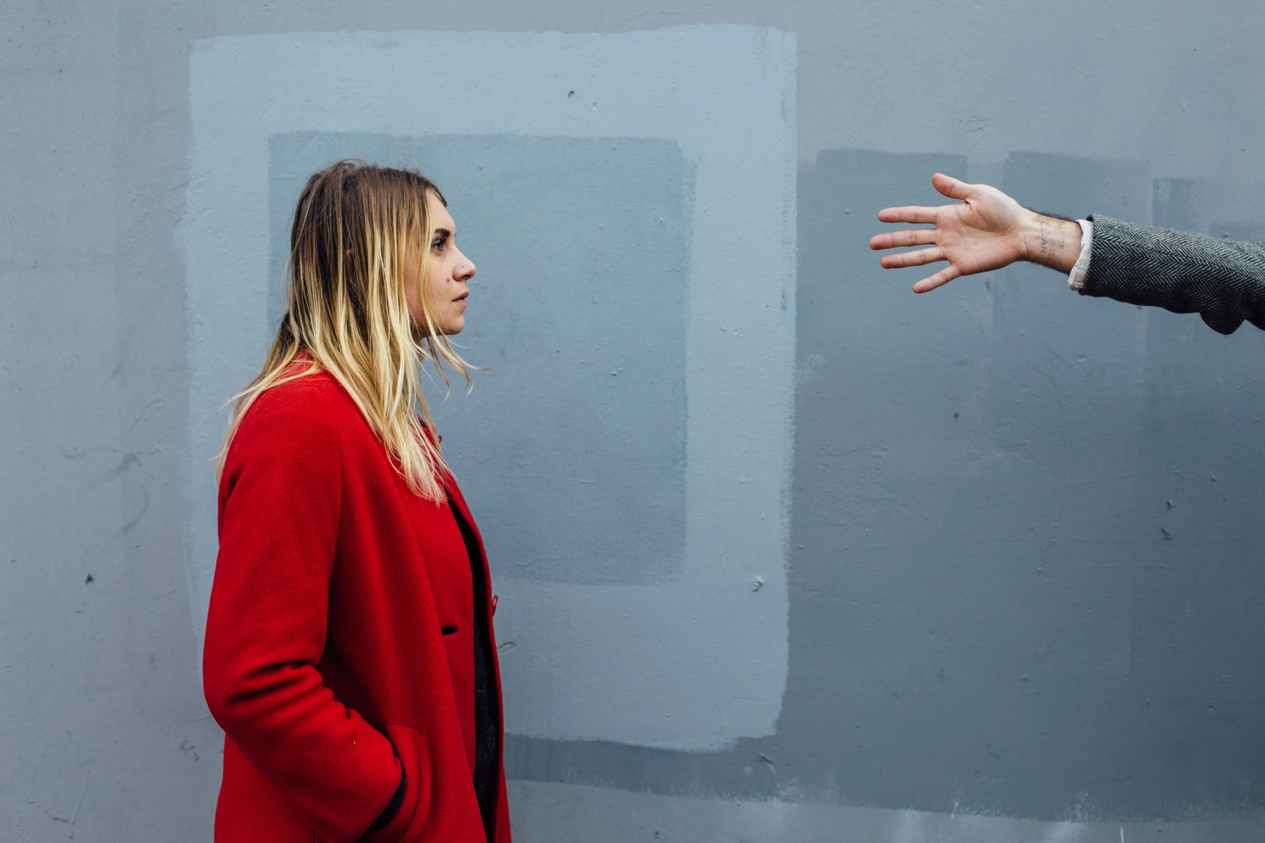 A woman in a red coat faces a person extending their arm with an outstretched hand, against a gray background.