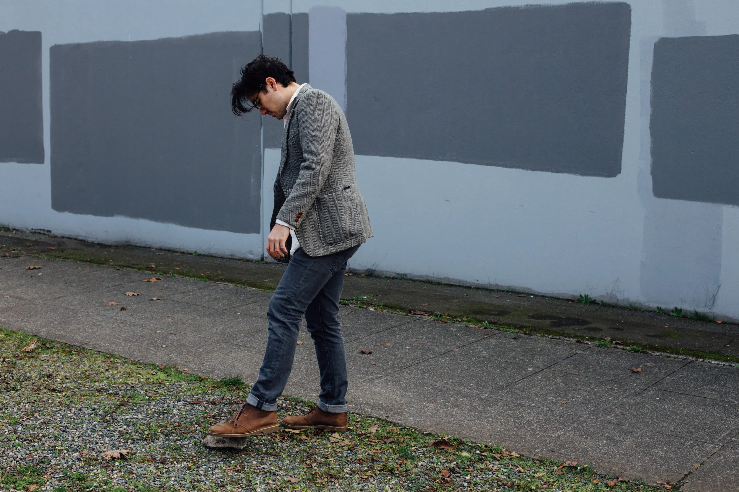 A man in a gray blazer, jeans, and brown boots standing with his head bowed, looking down at the uneven ground beside a sidewalk in front of a wall with painted gray rectangles.