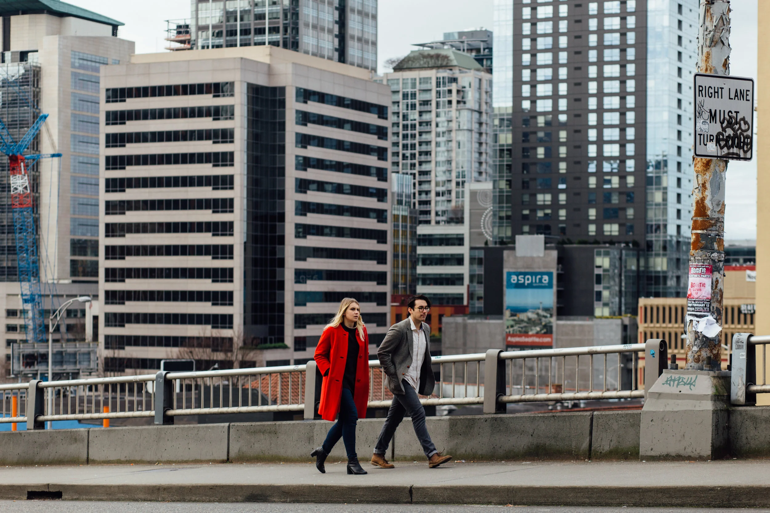 A woman in a red coat and a man in a gray jacket walk across a city street with tall office buildings in the background. A rusty pole with a sign and some posters is on the right side.