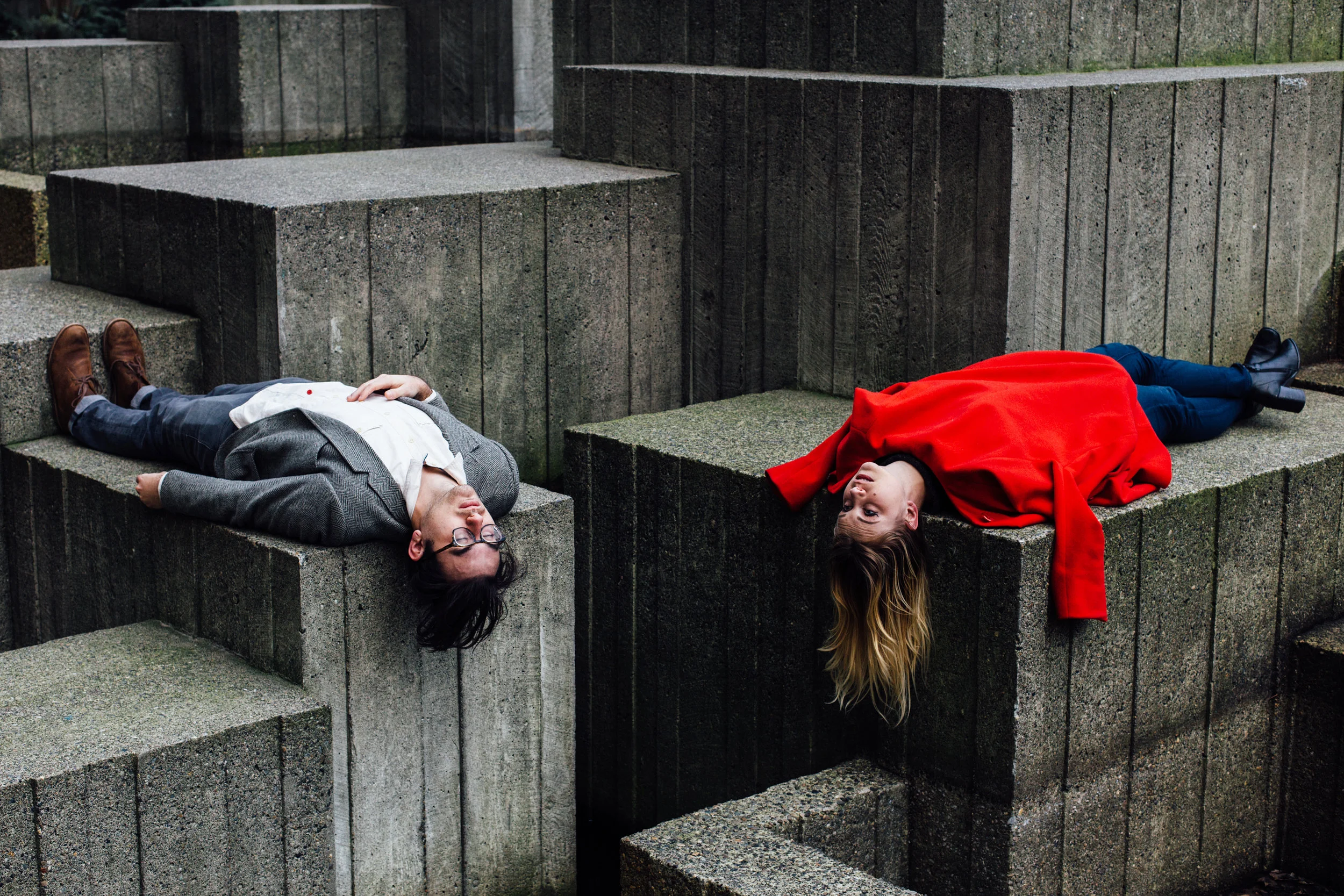 Two people lying on large concrete blocks outdoors, one man on the left in a gray suit and glasses, and one woman on the right in a red jacket, facing up with their heads hanging off the edges of the blocks.