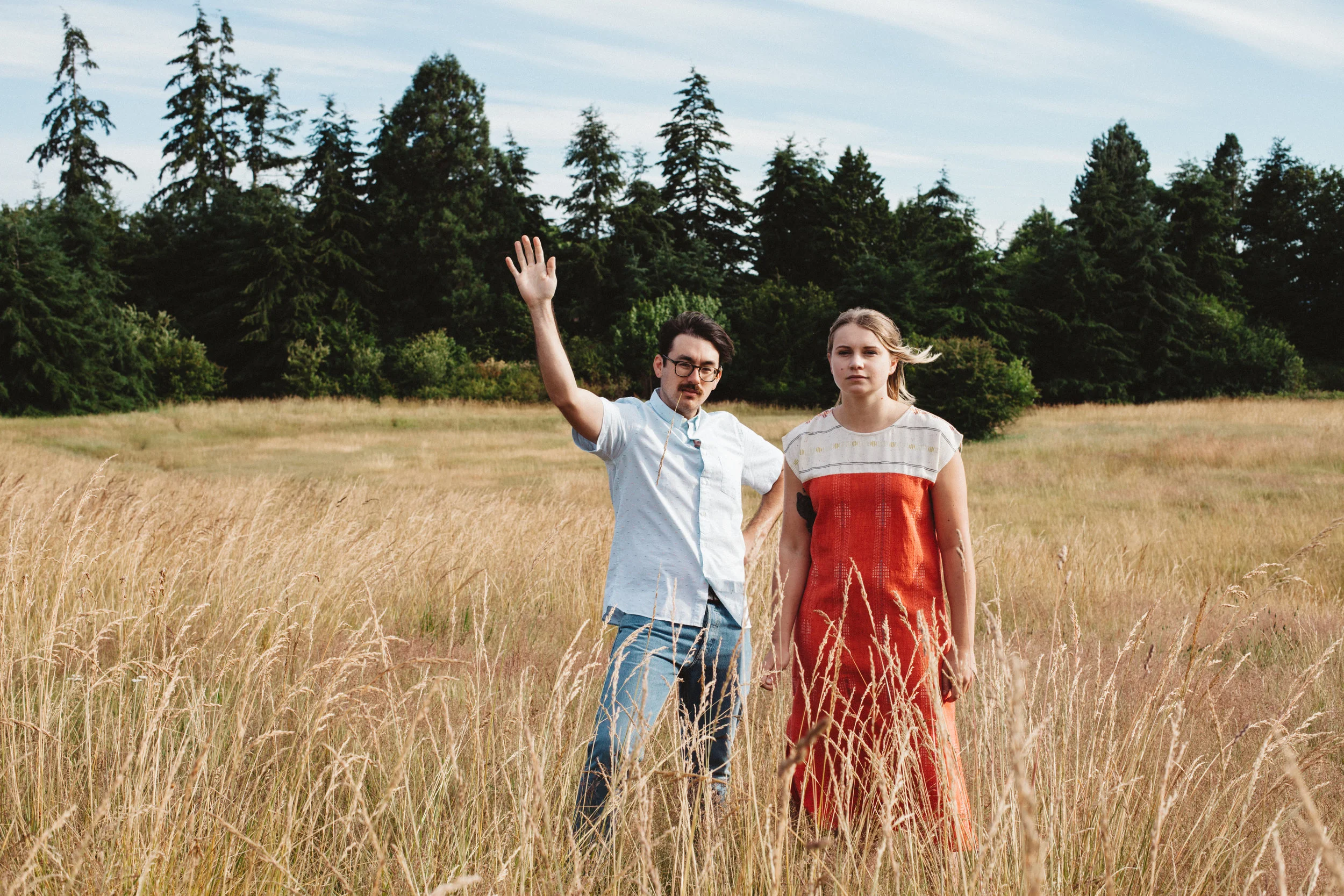 A man and a woman standing in a grassy field with trees in the background, the man waving his hand.