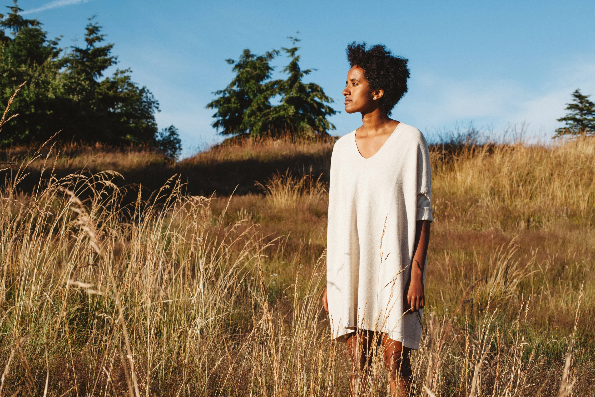 A woman with dark skin and natural curly hair stands in a field of tall golden grass, wearing a loose white dress, looking to the right against a backdrop of clear blue sky and trees.