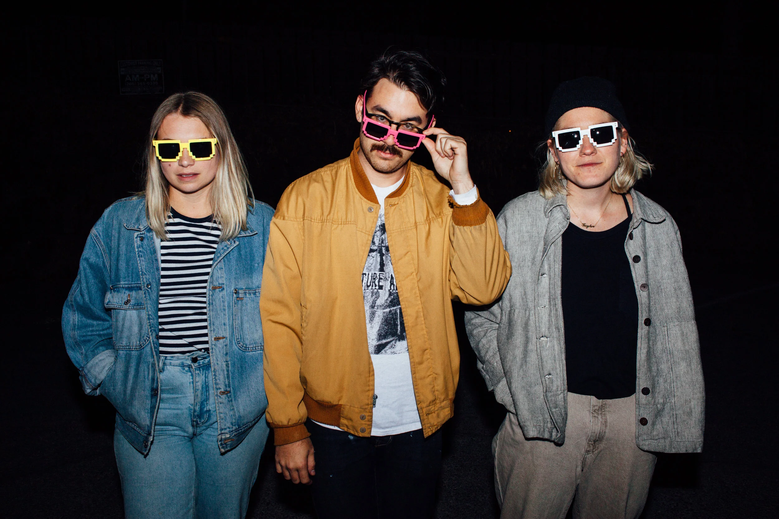 Three young adults standing together at night, wearing sunglasses and casual clothing, with a dark background.