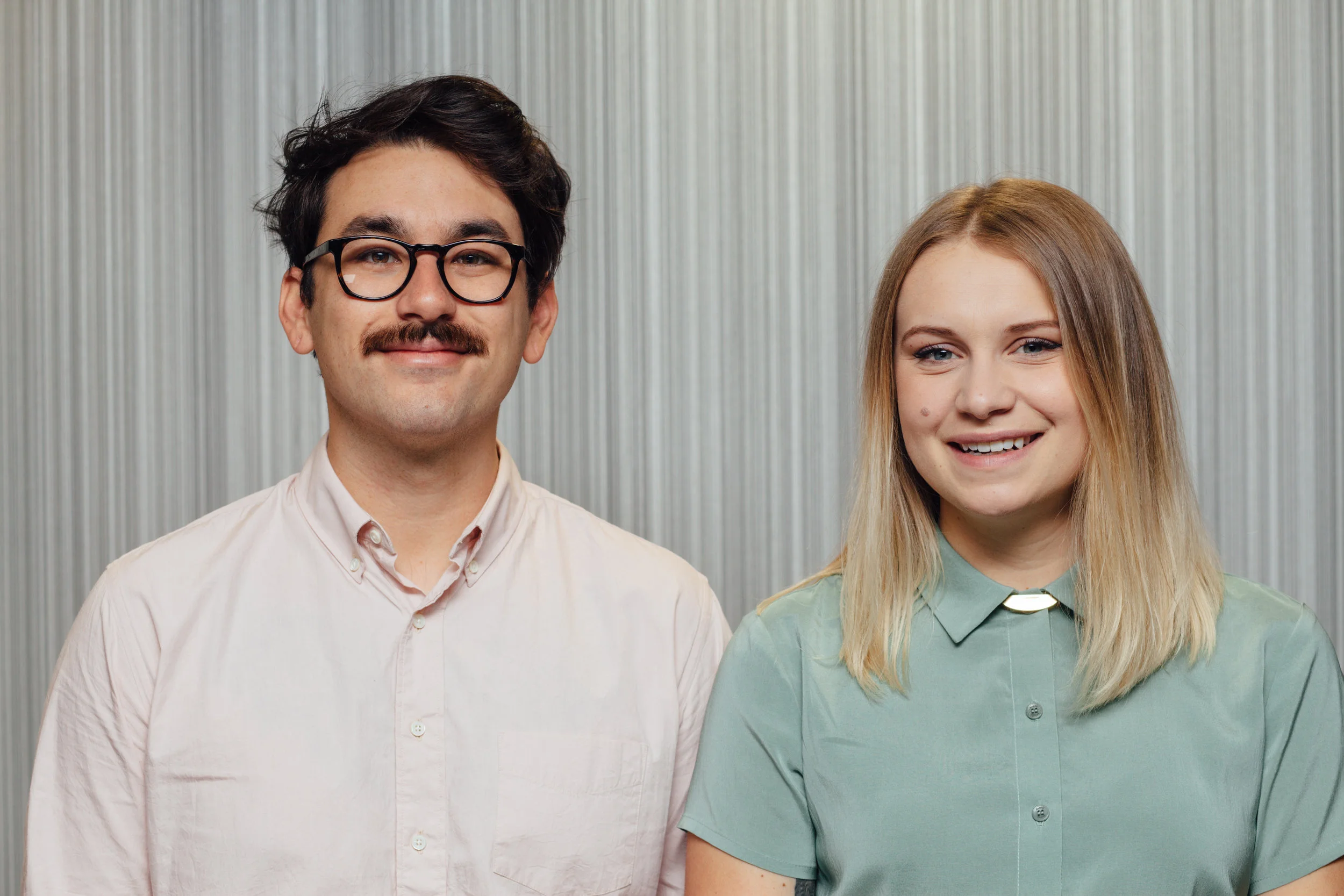 A young man with black hair, glasses, and a mustache smiling, standing next to a young woman with blonde hair and a light green shirt, both posing against a metallic background.