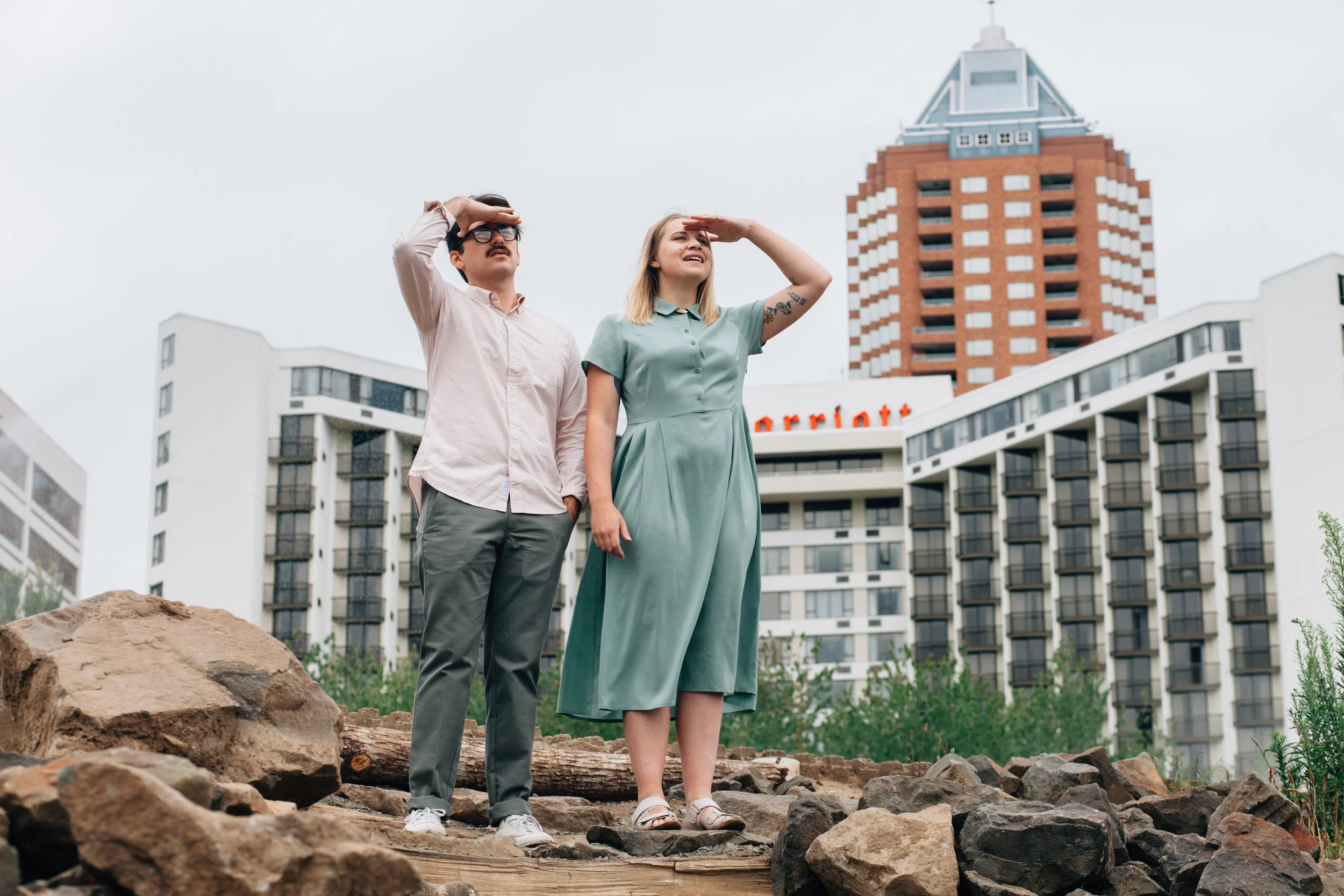 A man and woman standing outdoors on rocky terrain, looking into the distance with their hands shielding their eyes. In the background are modern buildings and a tall red-brick skyscraper under an overcast sky.