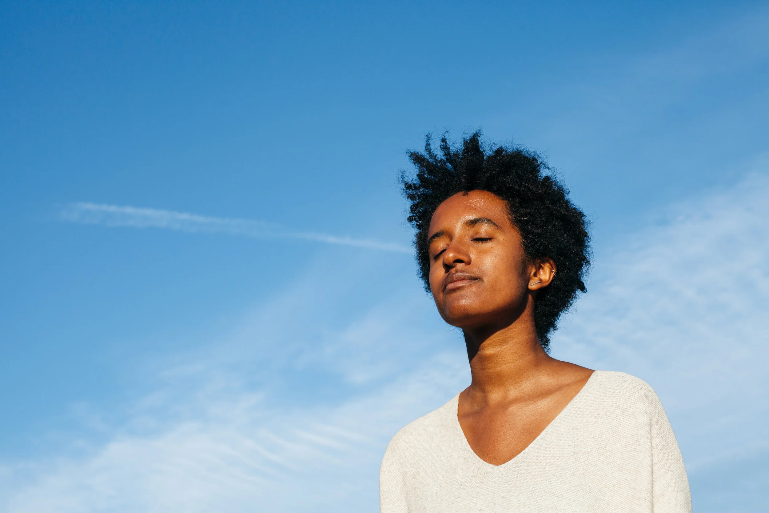 A woman with closed eyes and a peaceful expression stands outdoors under a blue sky.