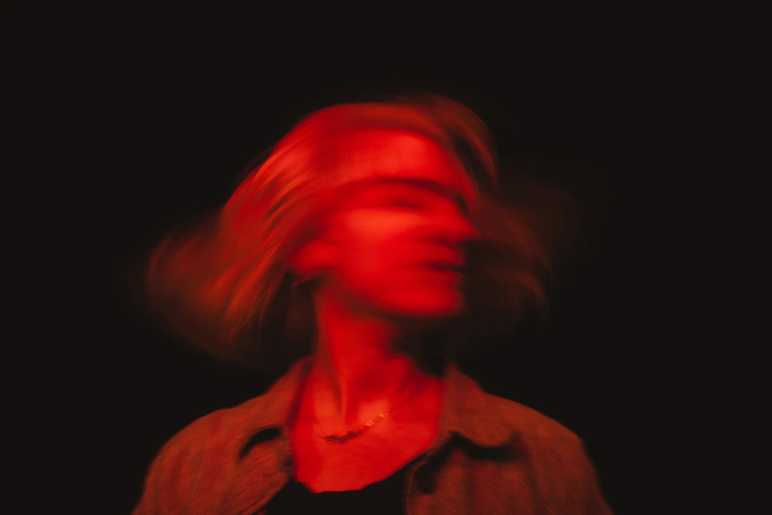 A person with wavy hair moving quickly, creating a blurred effect, with red lighting in a dark background.