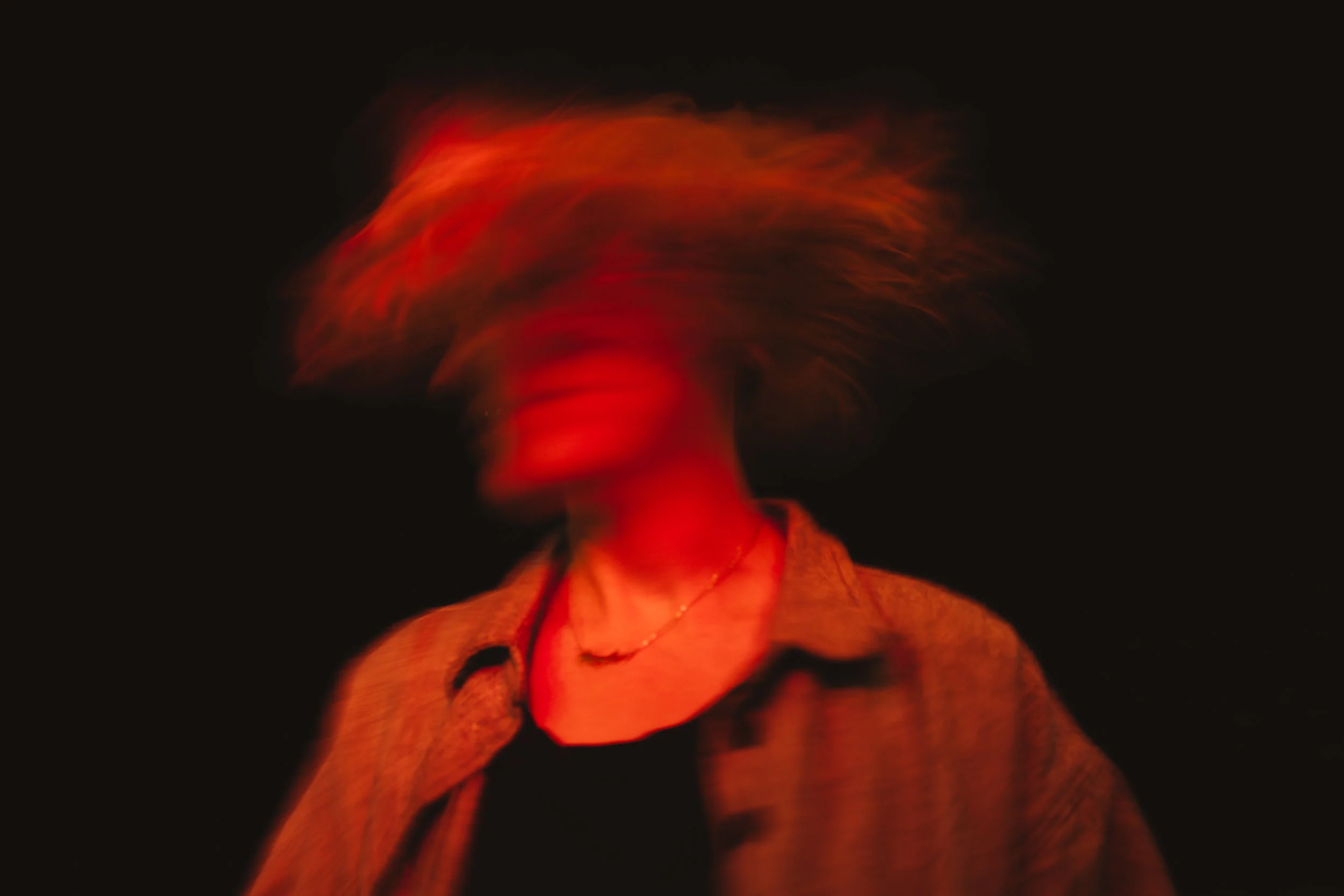 Blurry portrait of a woman with blonde hair, wearing a brown jacket and black shirt, against a black background, illuminated with red light.