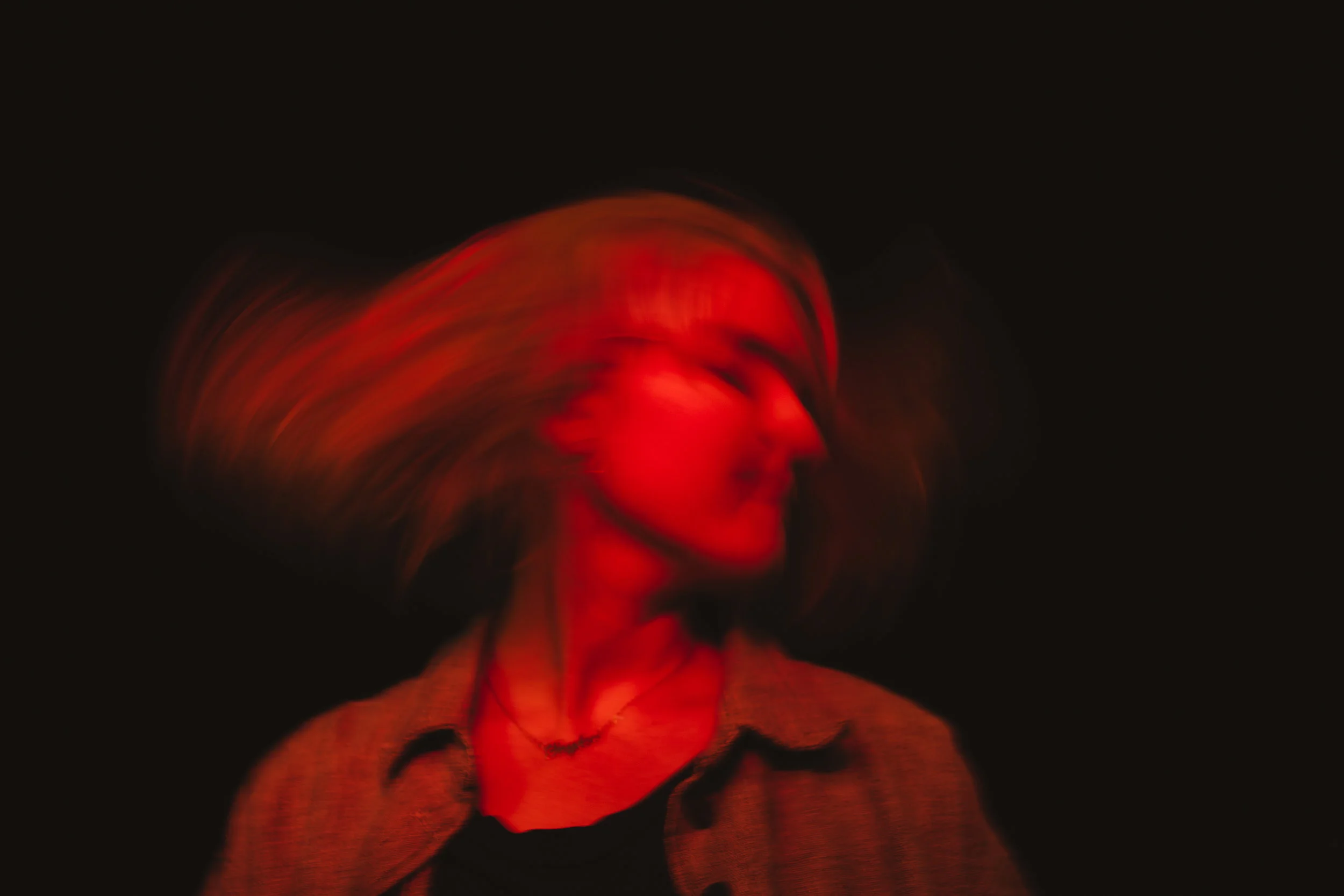 A person with shoulder-length hair is moving their head rapidly, causing a blurred effect, illuminated by red lighting in a dark environment.