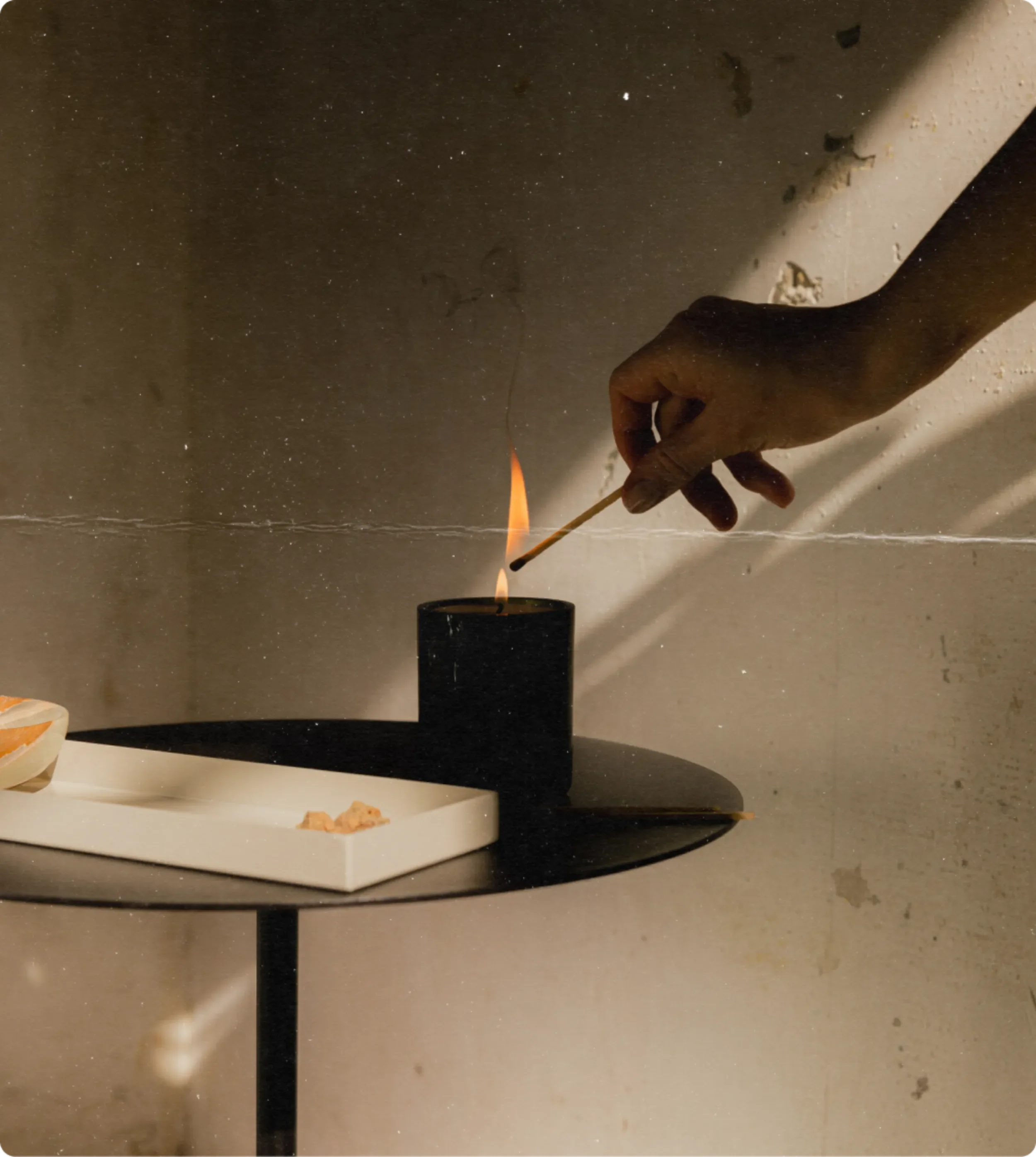 Person holding a lit matchstick near a small fire or flame, with a black object in a white dish on a table in front, against a gray background.
