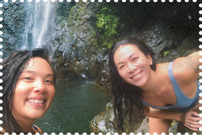 Two women smiling and taking a selfie in front of a waterfall in a forested area.