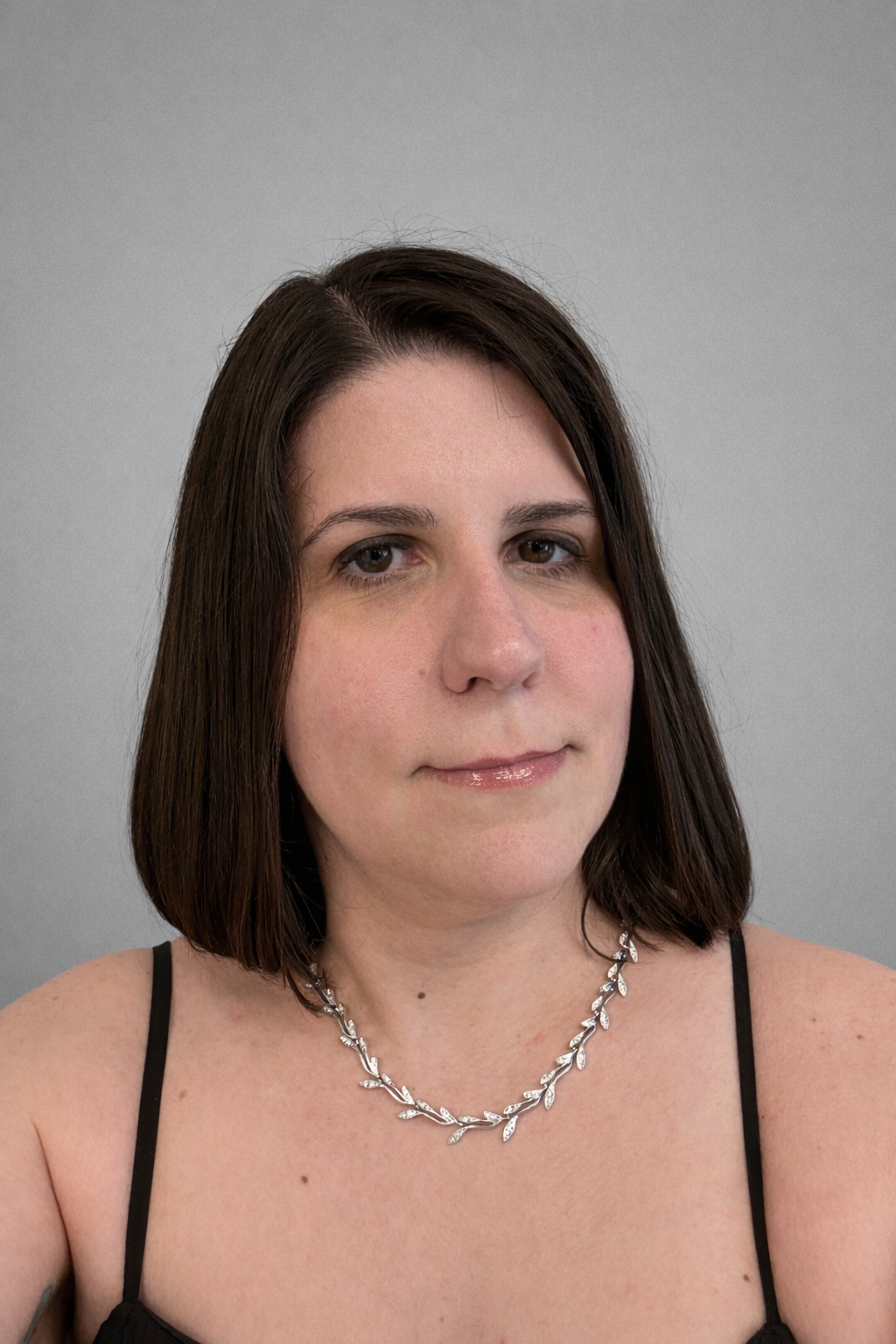 A woman with shoulder-length brown hair, wearing a dress and a silver necklace with leaves, posing against a plain gray background.
