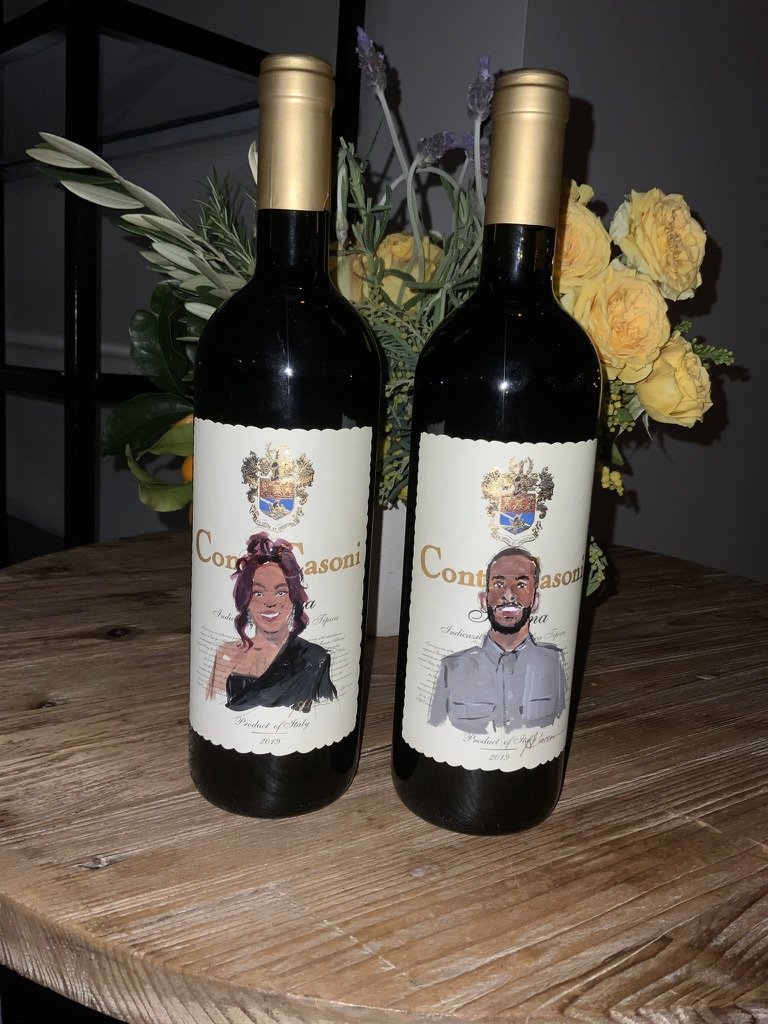 Two bottles of wine with custom labels featuring illustrated portraits of a woman and a man, placed on a wooden table with a flower arrangement in the background.
