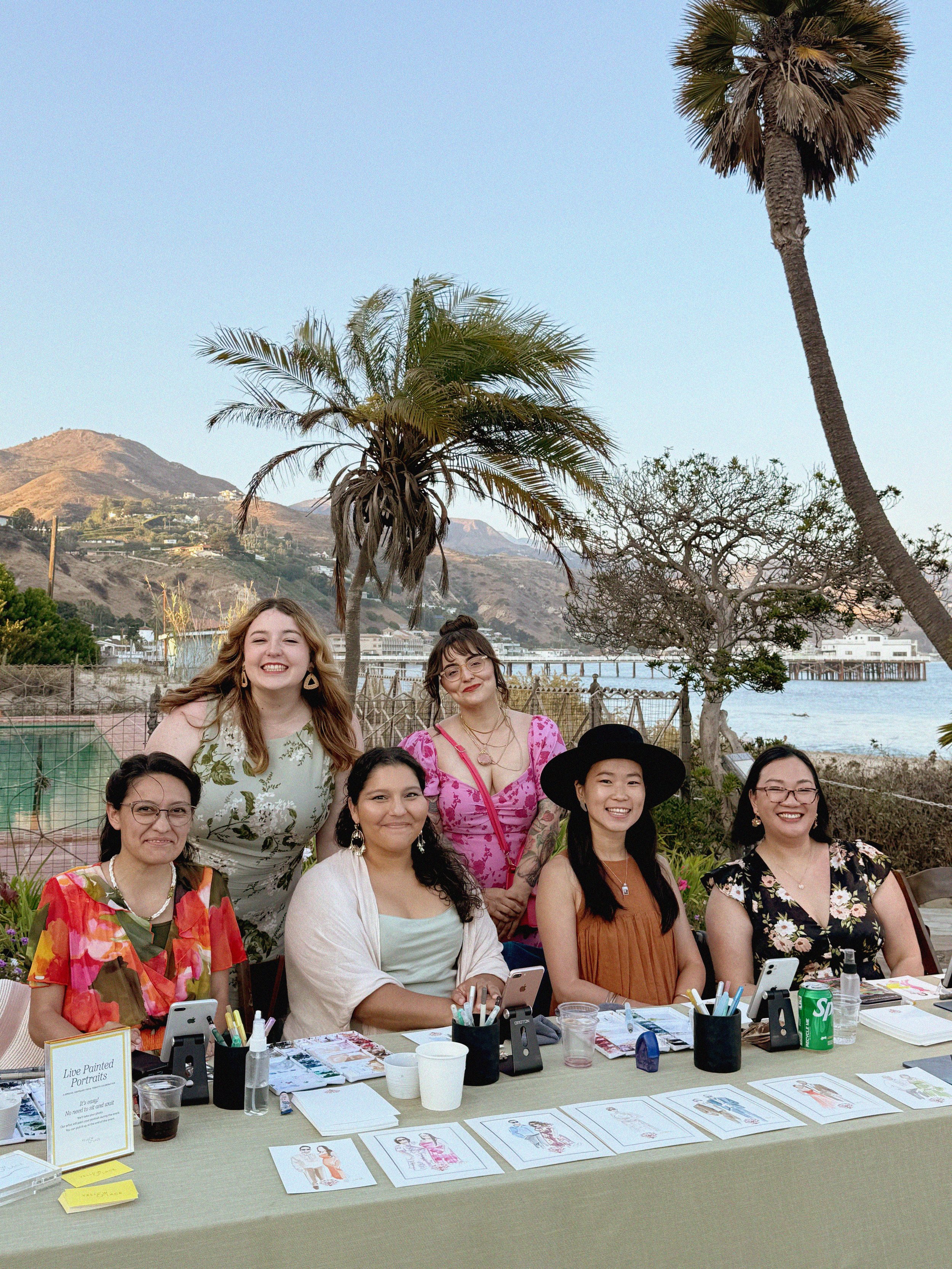 Group of seven women smiling around a table outdoors with palm trees, a hill, and water in the background.