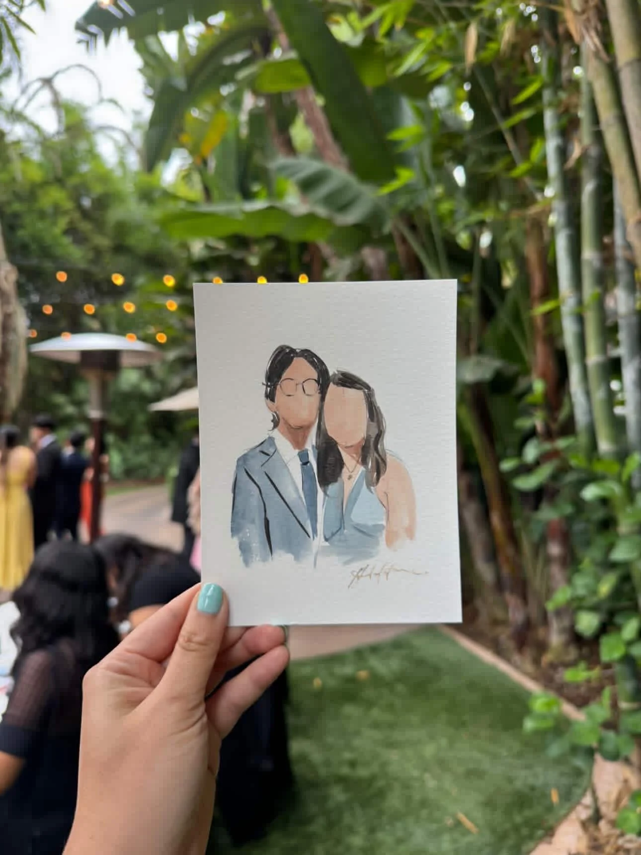 Hand holding a watercolor portrait of a couple, with a man wearing glasses and a suit, and a woman with shoulder-length hair, at an outdoor gathering with lush greenery and string lights in the background.