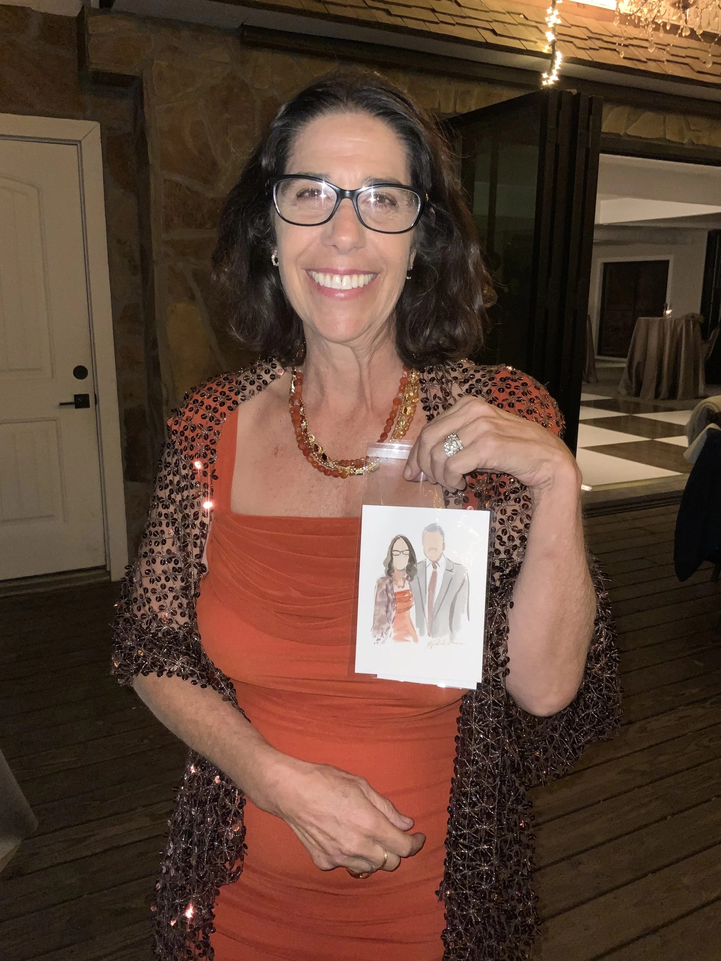 A woman smiling and holding a cartoon caricature of herself and a man. She has shoulder-length dark hair, glasses, and is wearing an orange dress with a beaded shawl and necklace.