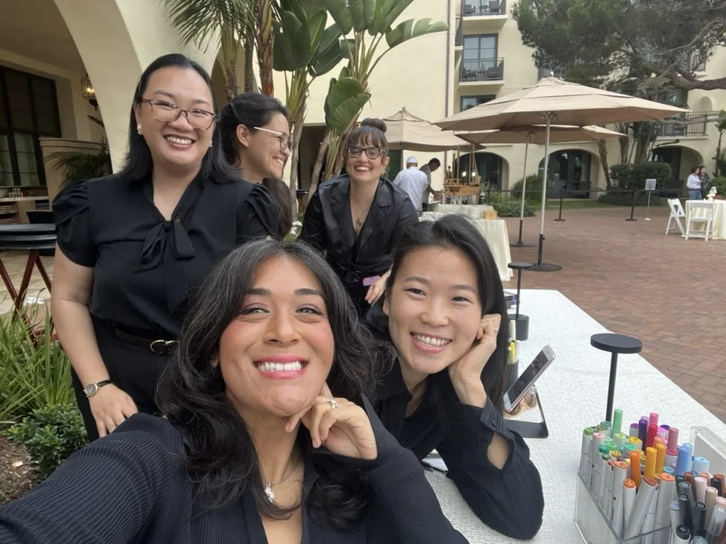 Group of five women smiling and taking a selfie outdoors at a patio with umbrellas, plants, and a multi-story building in the background.
