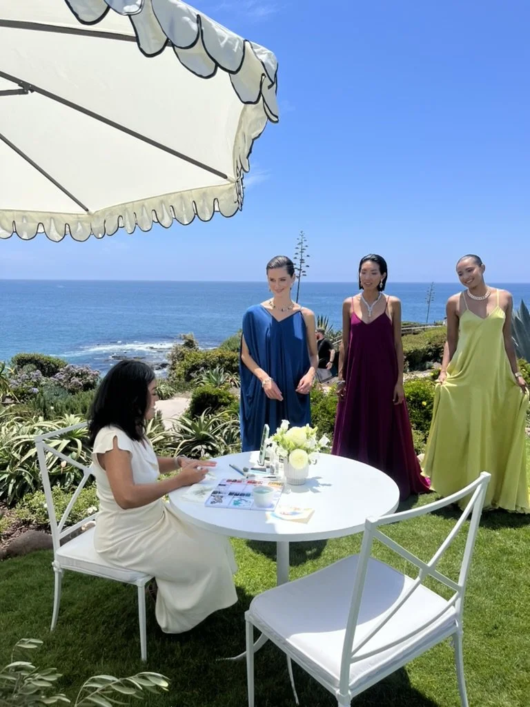 A woman in a white dress sitting at a white table outdoors under a large umbrella with a scenic ocean view, while three women in colorful dresses stand nearby on the grass.