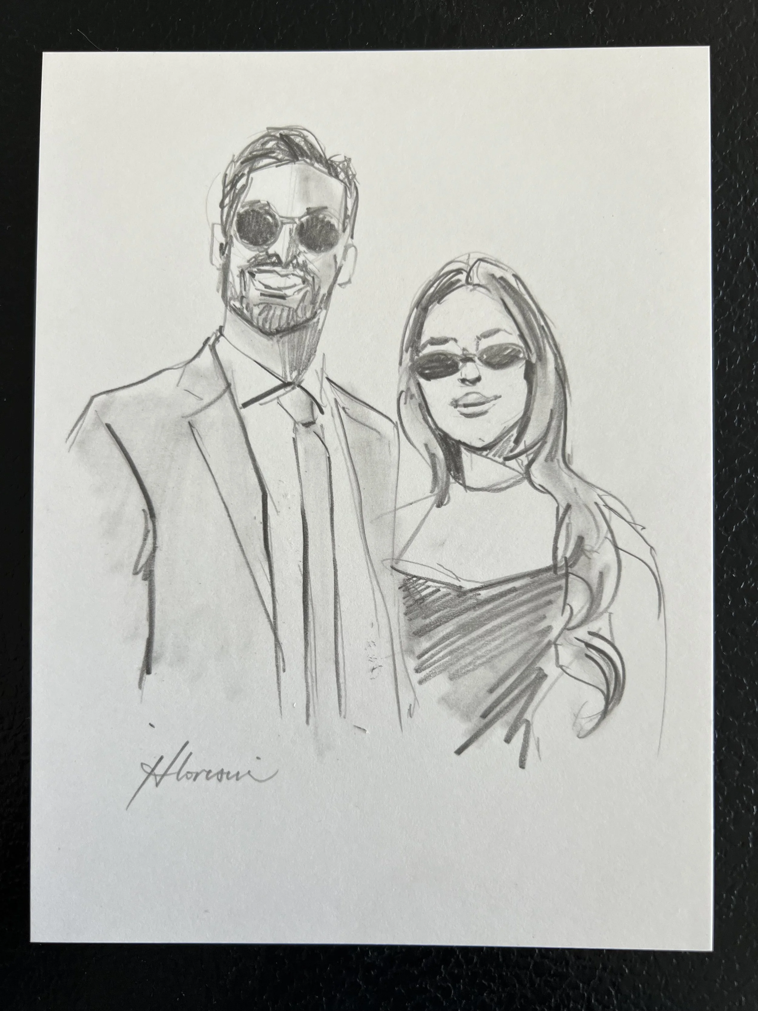 A sketch of a man and woman, both wearing sunglasses. The man has facial hair and is dressed in a suit with a tie, while the woman has long hair and is wearing a top. The drawing is signed with the name 'Horaci'.