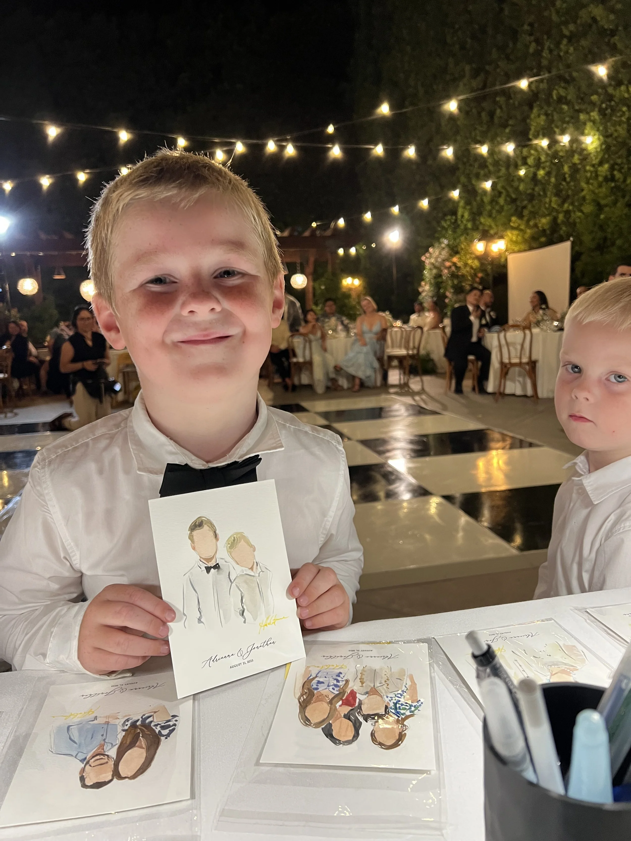 A young boy wearing a white dress shirt and black bow tie at a nighttime outdoor celebration, holding a painting of a faceless couple in formal attire, with decorated tables and string lights in the background.