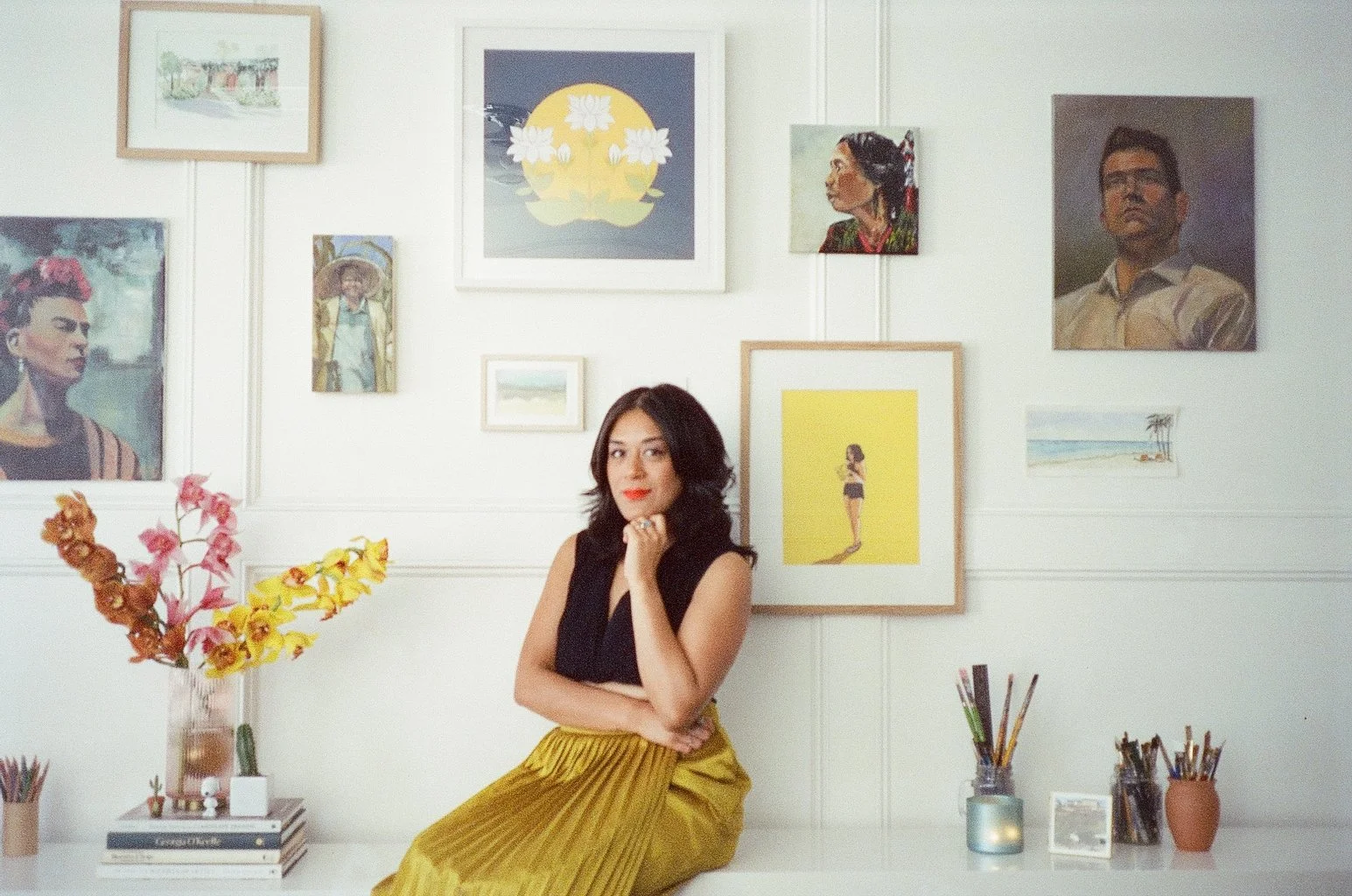 A woman with dark hair and a red lipstick sitting on a white surface in front of a gallery wall with various framed artwork, including portraits, landscapes, and abstract pieces. She is wearing a black sleeveless top and a gold pleated skirt, with her left hand resting on her chin.