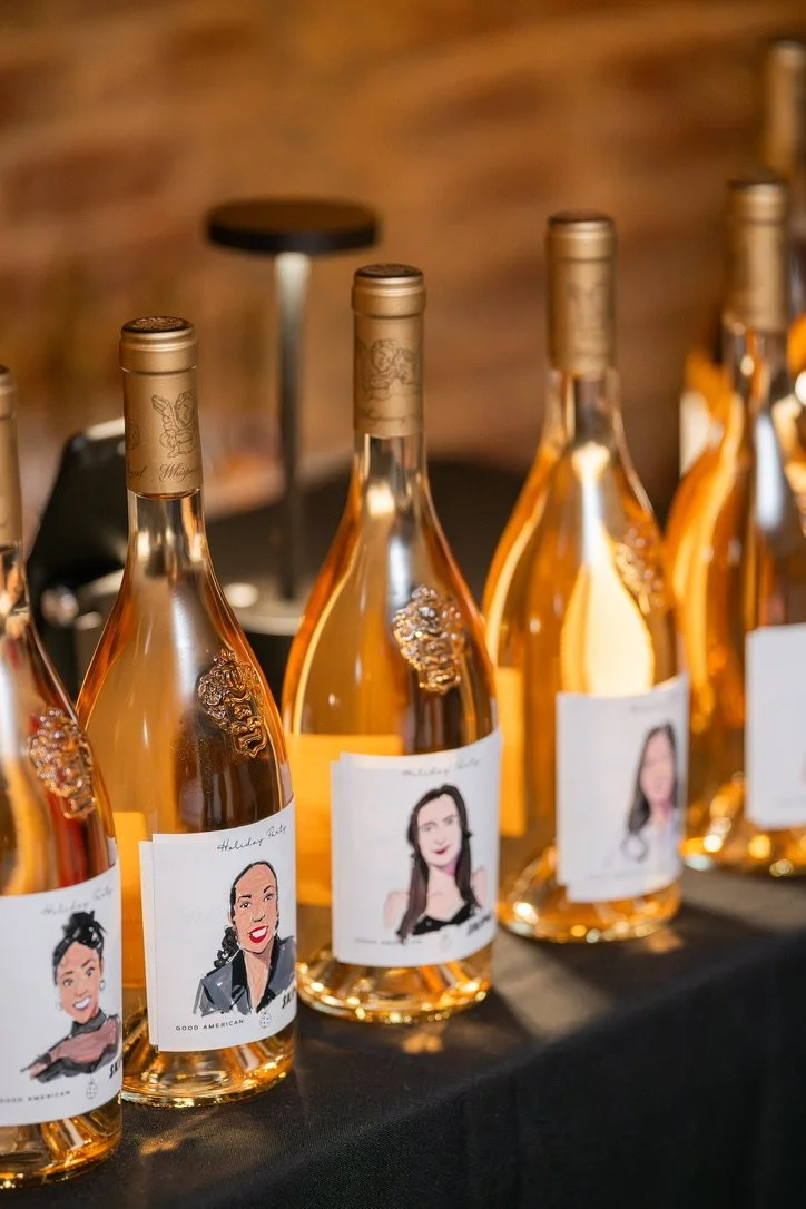 Rose wine bottles with personalized labels featuring cartoon portraits of women, arranged on a black table.