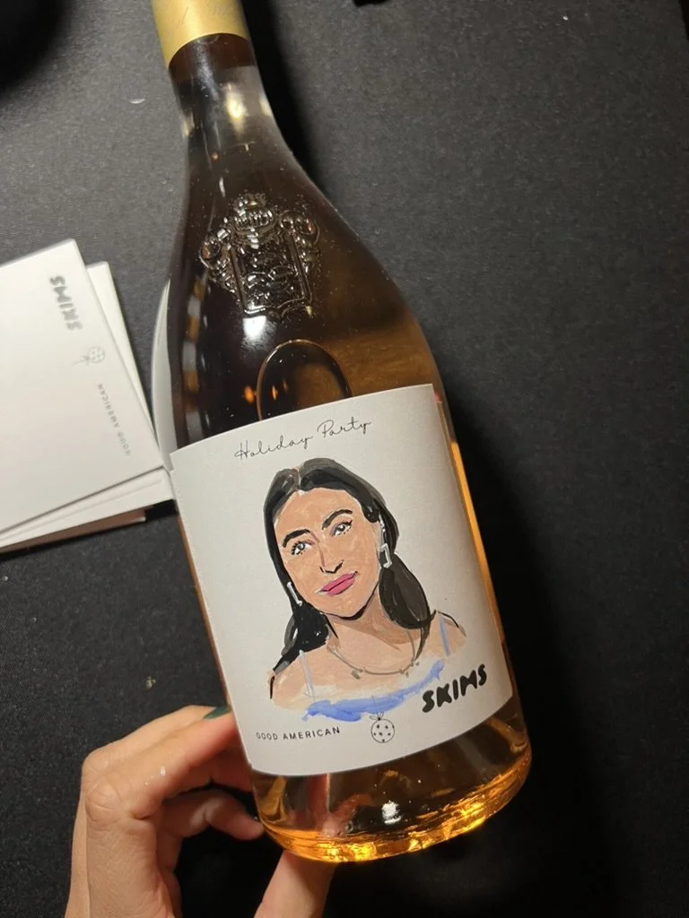 A hand holding a bottle of wine with a custom label featuring a cartoon portrait of a woman with dark hair, earrings, and makeup. The label reads "Holiday Party" and has the words "Good American" and "Skims".