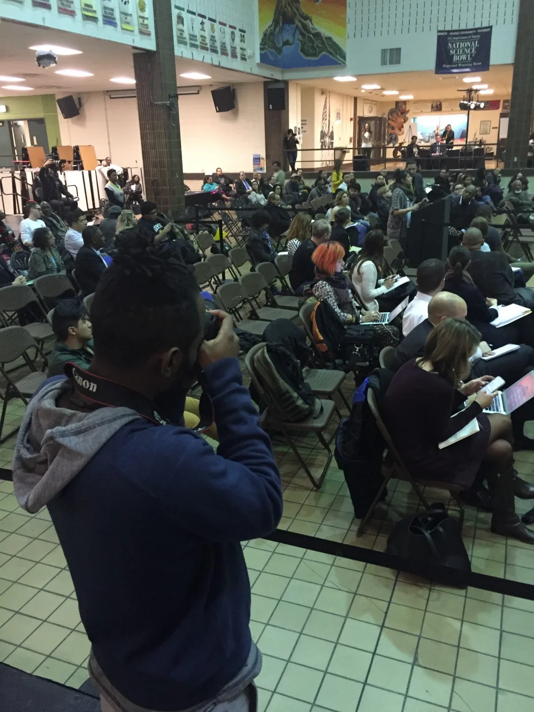 A former youth mentee from Free Spirit Media’s Real Chi Youth program (foreground, with camera)) covers a Police Accountability Task Force meeting at Benito Juarez High School.