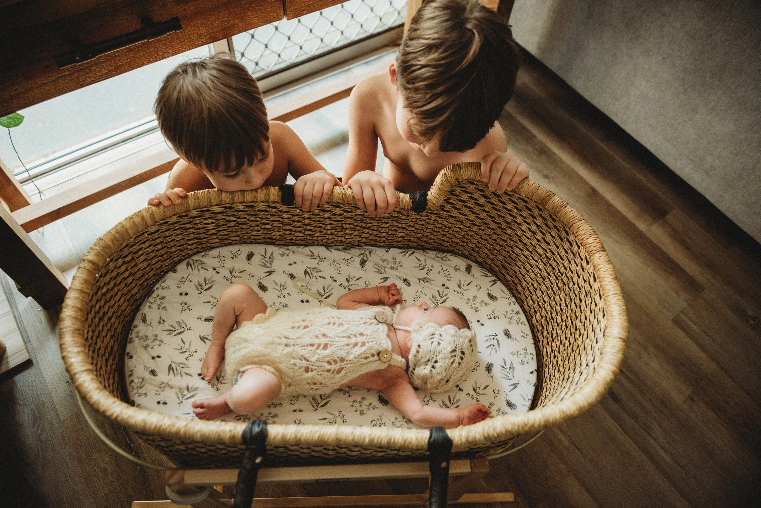 Two young Darwin boys looking down into a bassinet where a newborn baby is lying, wearing a knitted bonnet and outfit, in a room with wooden floors and a sliding door, lifestyle photography Darwin.