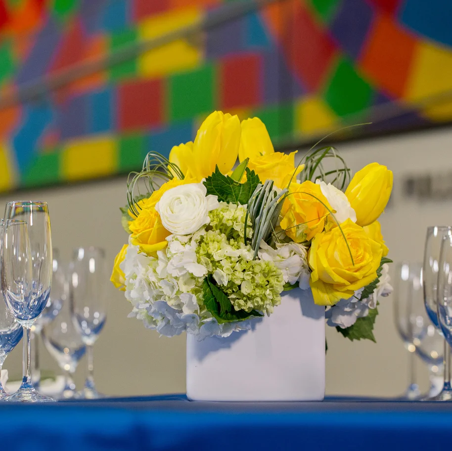 A white ceramic square vase is filled with white and mini green hydrangea and studded with yellow roses, spray roses, tulips and white ranunculus. Loops of bear grass complete the modern design. Image by Kendal J. Bush Photography