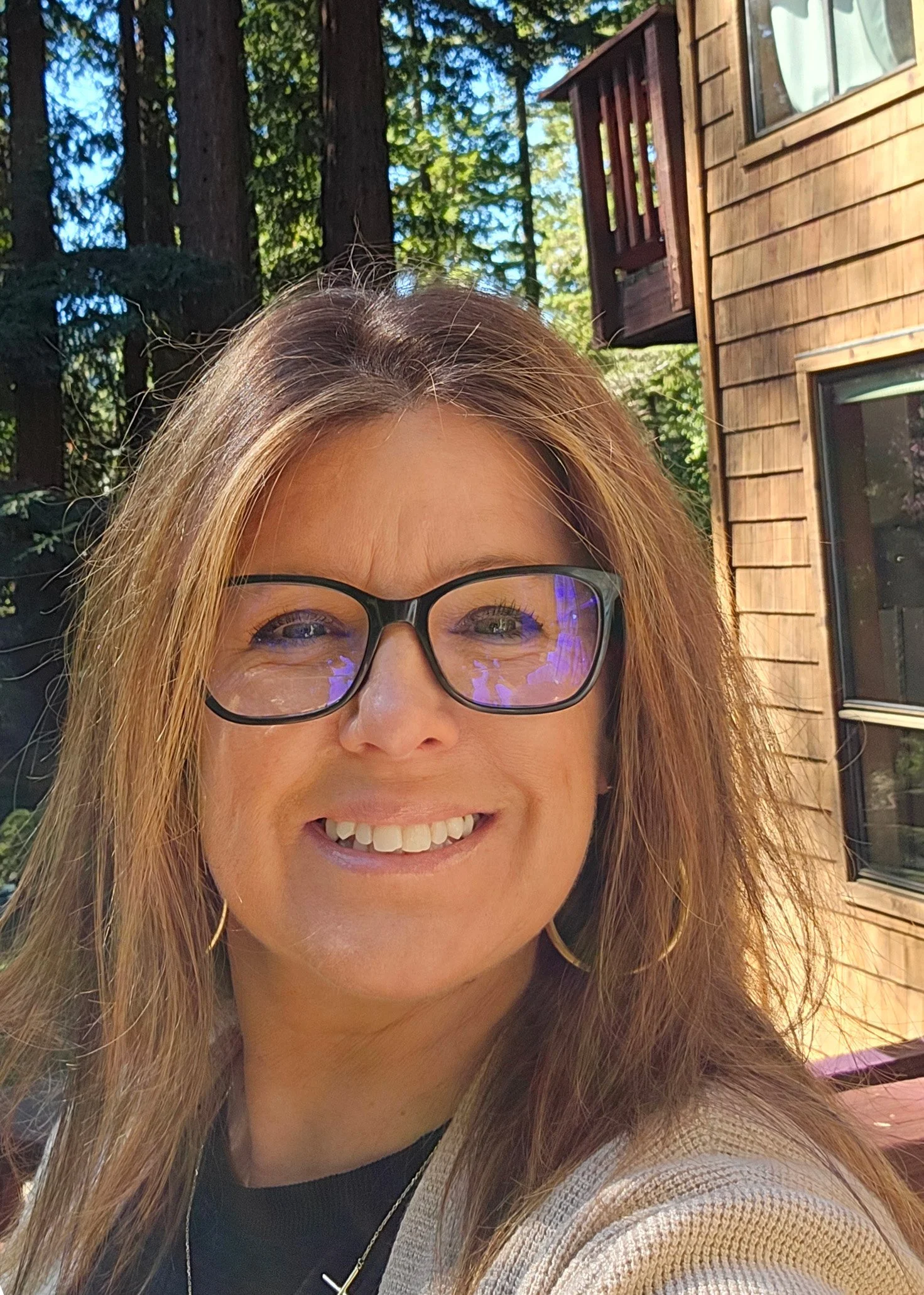 Smiling woman with shoulder-length hair and large hoop earrings standing outdoors in a lush green garden with trees and pink blossoms in the background.