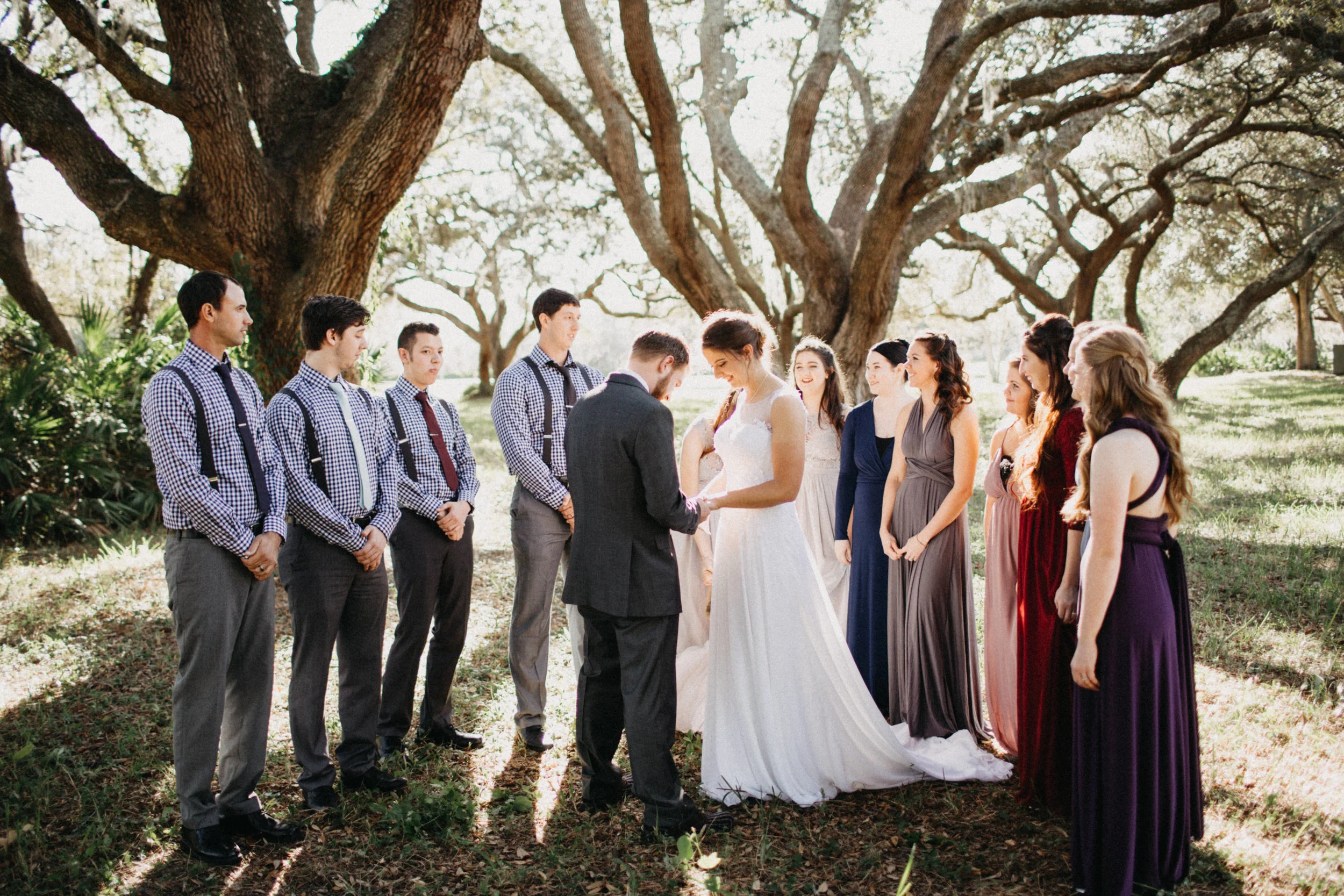 Photo of bridal party praying with bride and groom under trees.