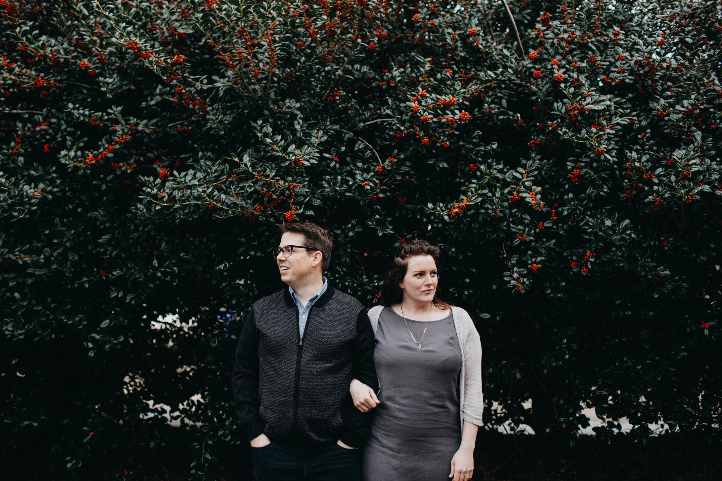 Couple standing together beside holly bushes.