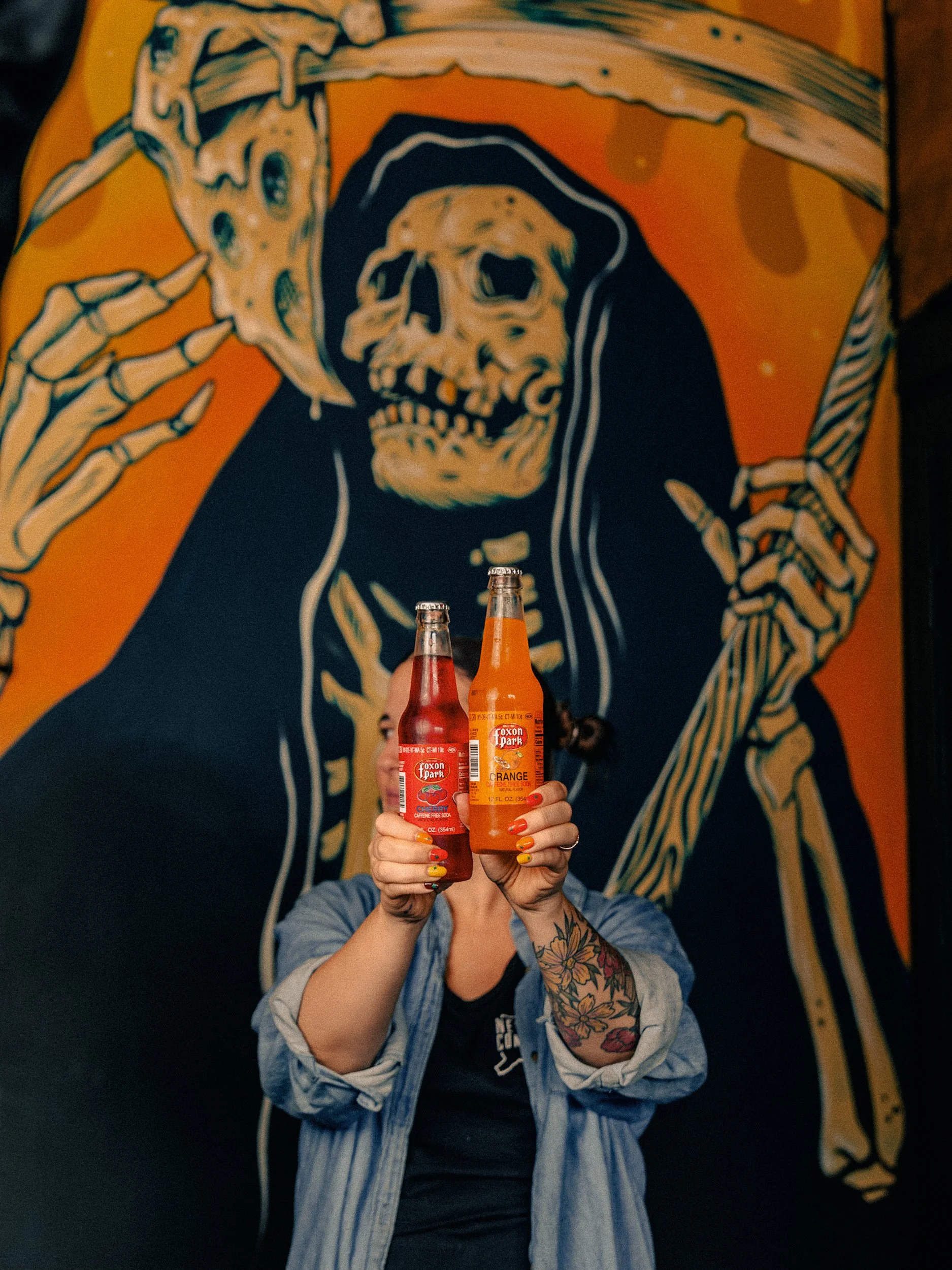 Owner Alexa holding two Foxon Park sodas in front of the Reaper mural at One6Three