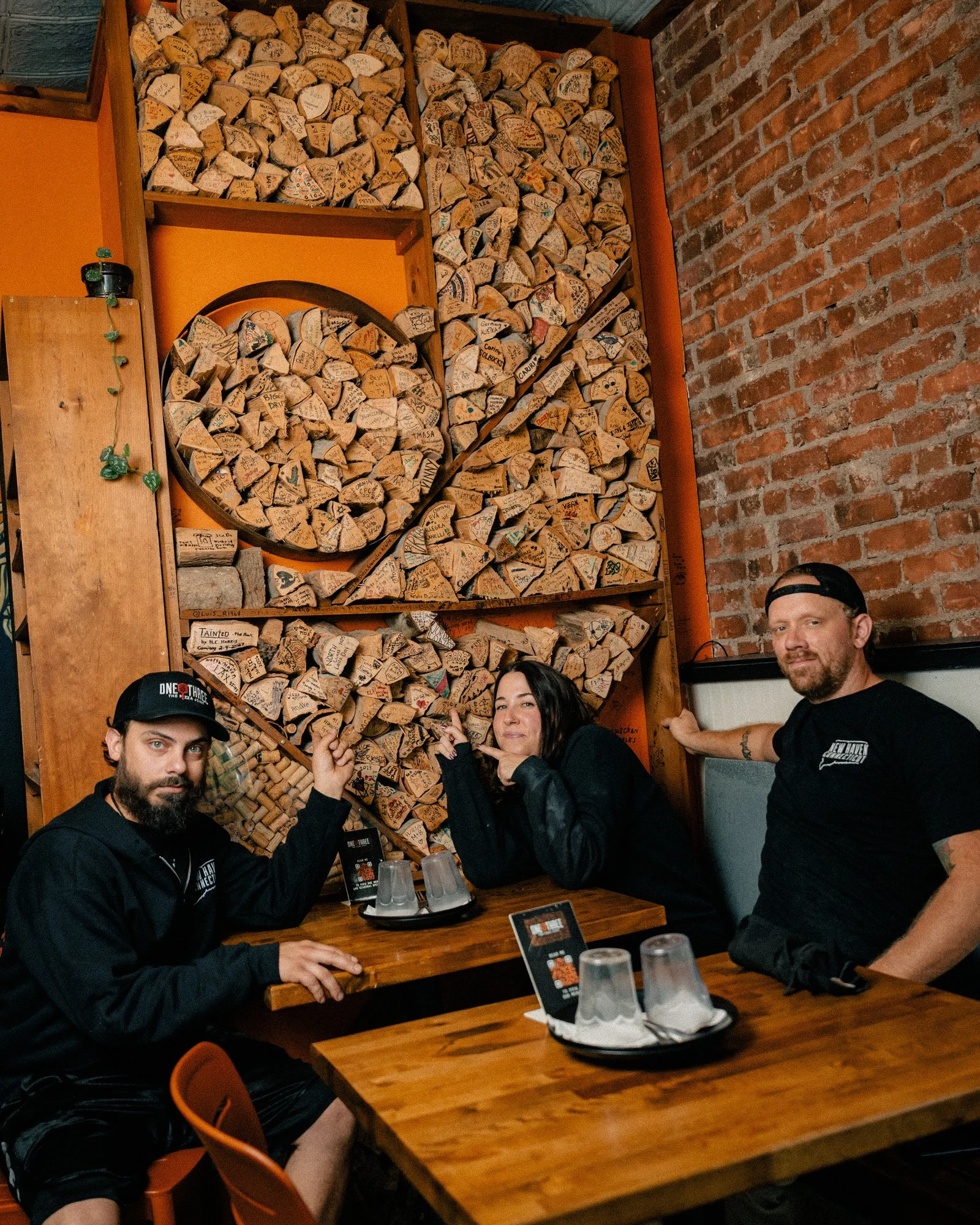 Owners Alexa and Andrew and pizza guy Chris at a table in front of the signed wood wall and brick at One6Three in East Rock
