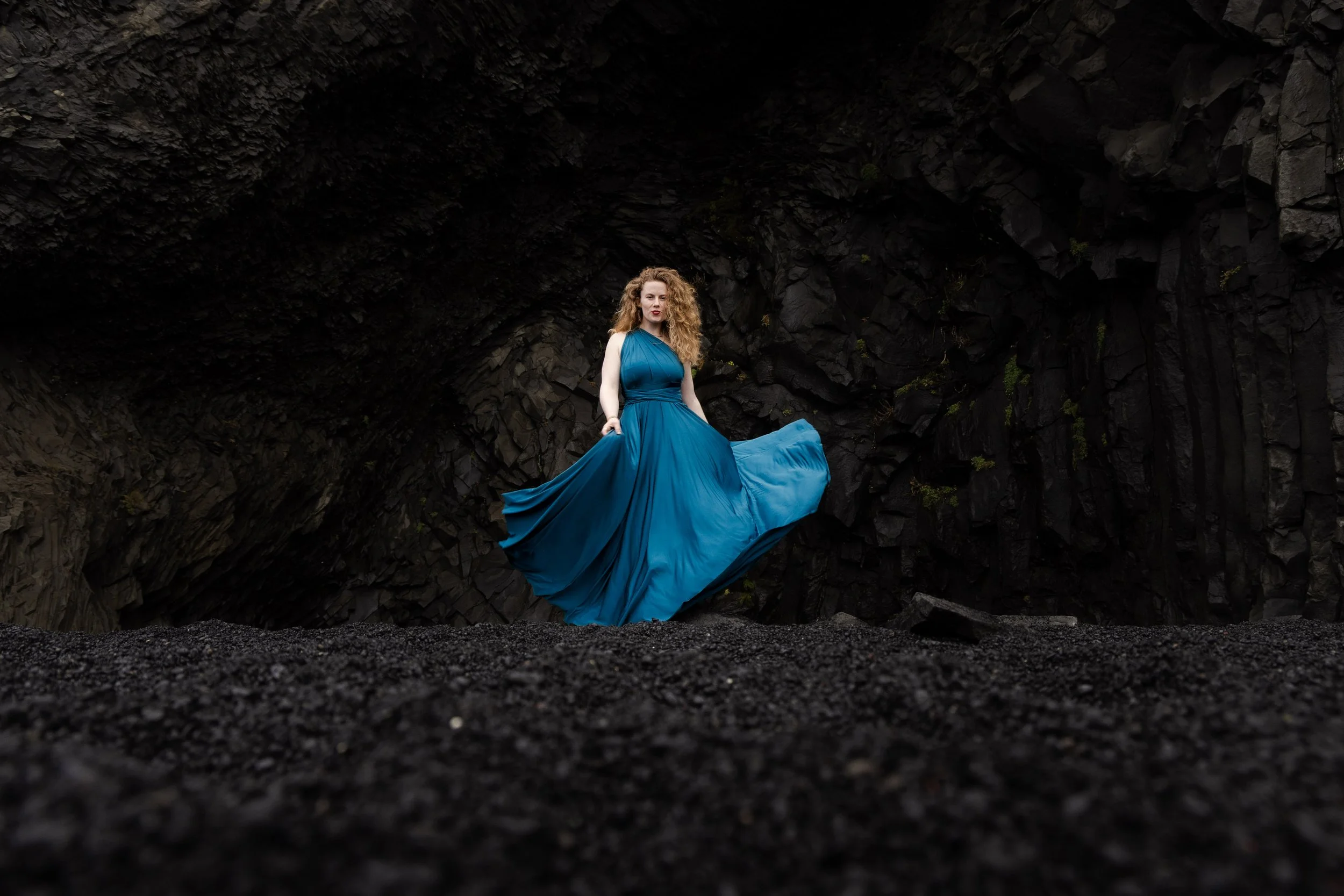 A woman with curly hair in a long, flowing blue dress standing in front of a dark rocky cave background. Folklore and fairytales.