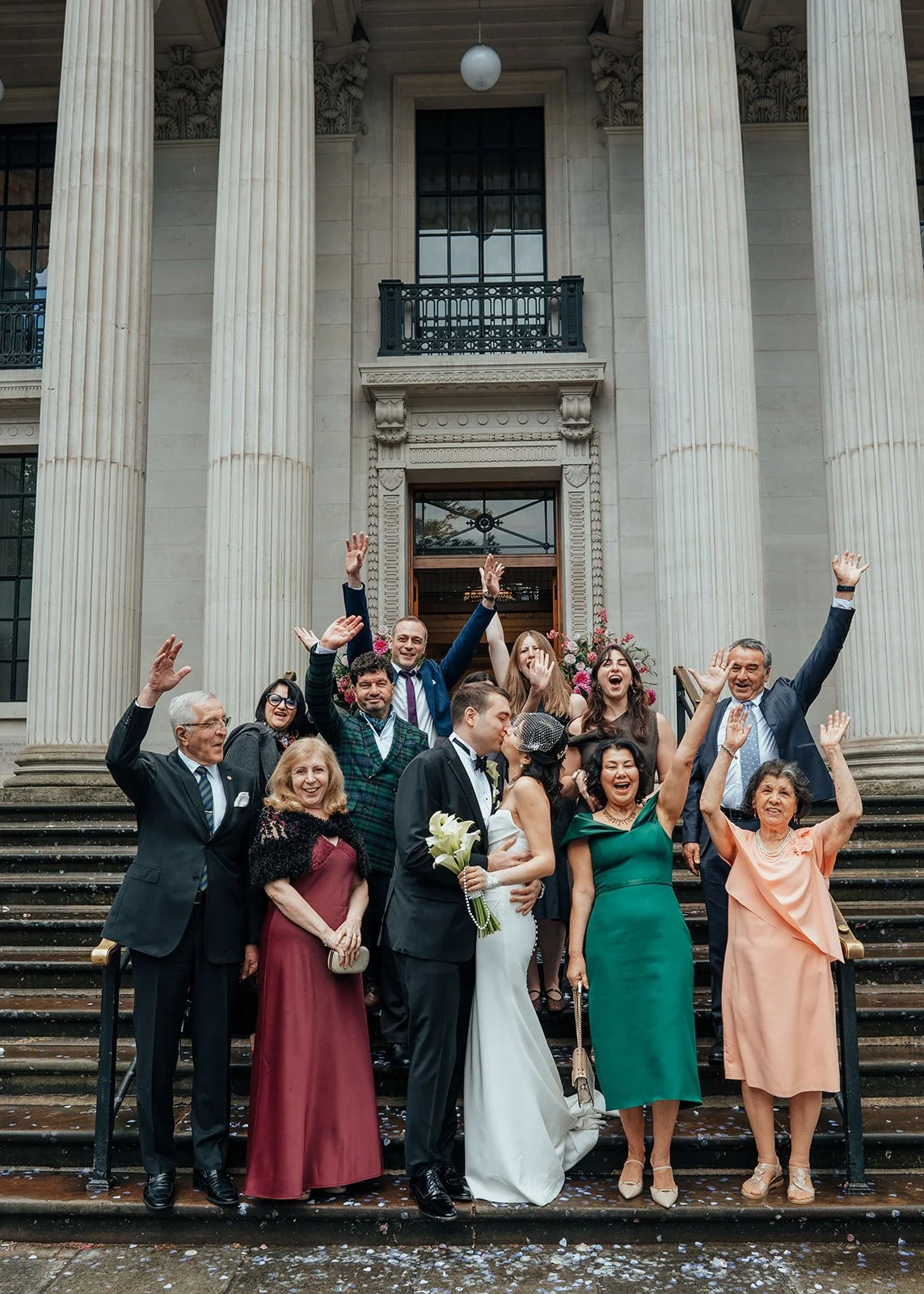 A newlywed couple kissing surrounded by their guests cheering outside Old Marylebone Town Hall
