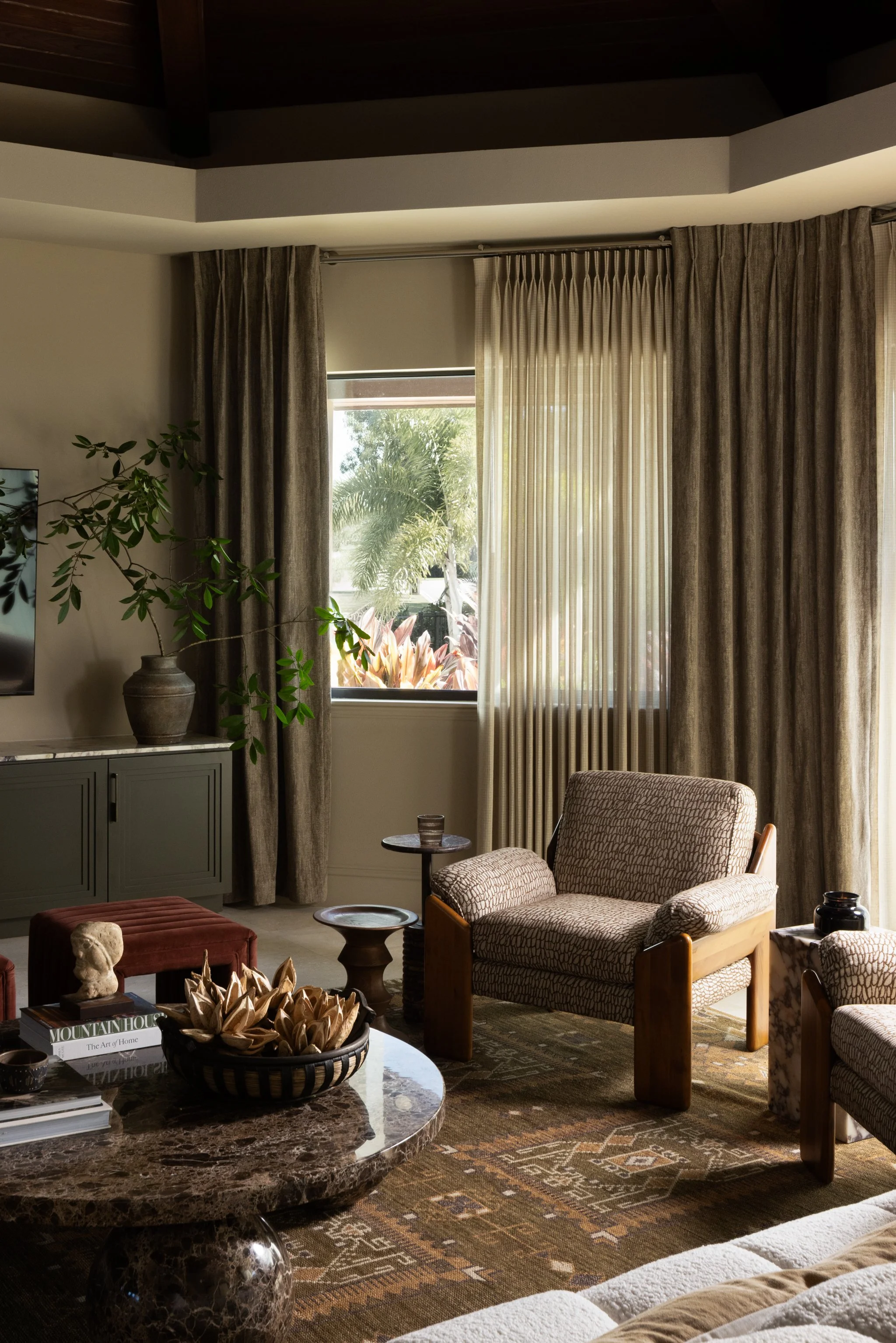 A cozy living room with a patterned armchair, side table, coffee table, and plants near a window with curtains, sunlight streaming in.