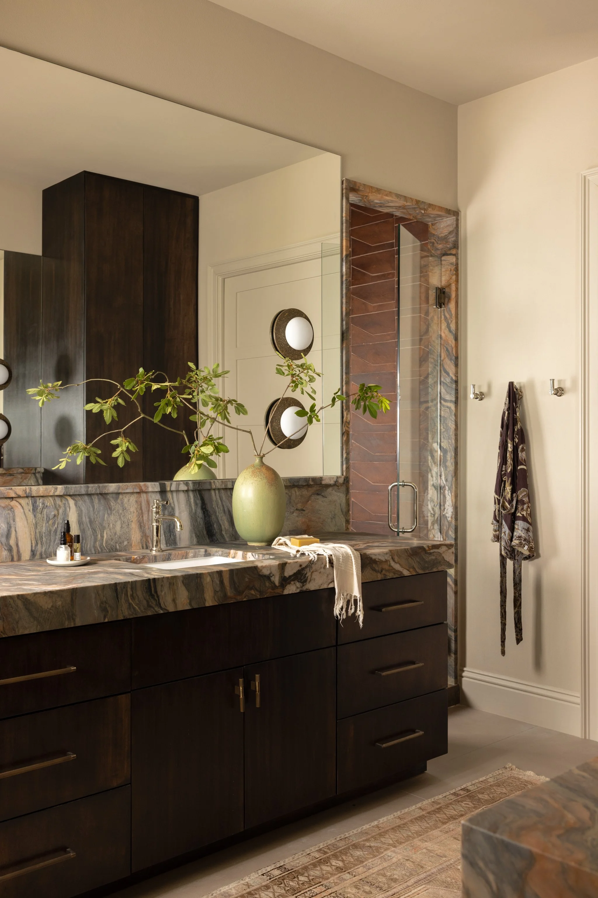 View of a bathroom vanity with a marble countertop, dark wood cabinetry, and a large mirror surrounded by three pairs of circular wall lights. A vase with greenery and a soap dispenser are on the countertop, with a rug on the floor.