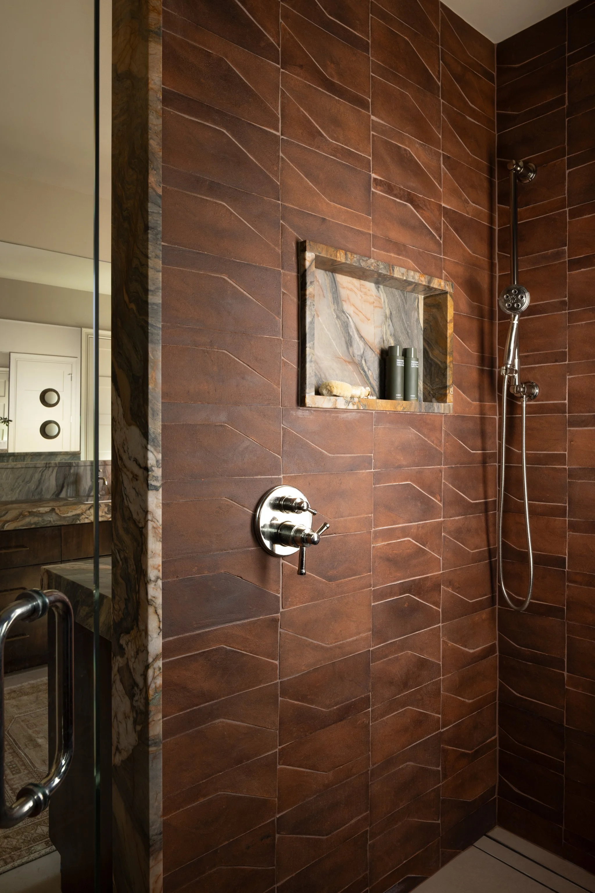 A modern shower area with rust terracotta tile walls, a built-in quartzite inset shelf with toiletries, a round polished nickel shower control, and a hand-held showerhead.