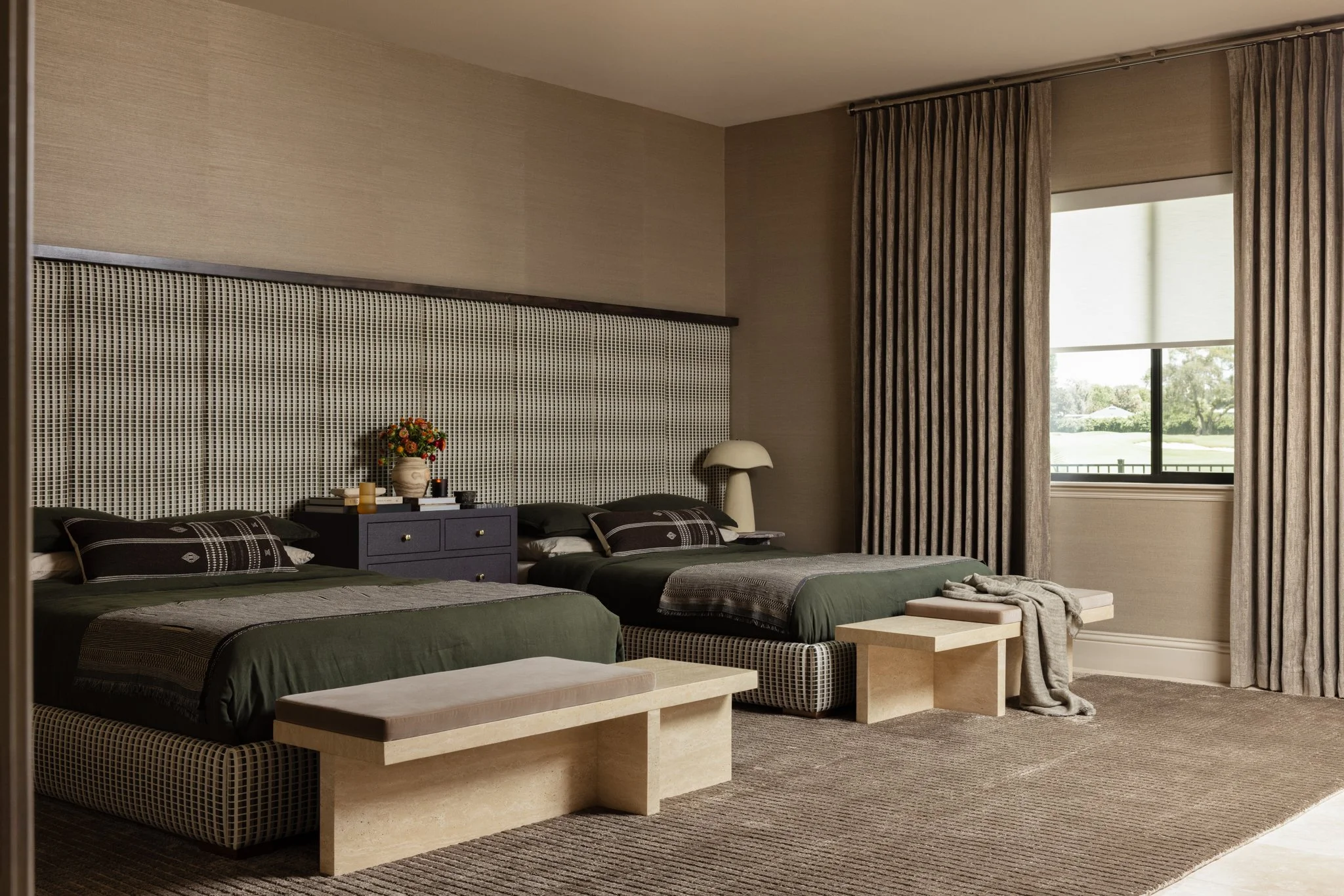 A master bedroom with two custom upholstered beds, a dark purple nightstand in between, decorated with a vase of flowers and books, and two matching cream-colored lamps on marble side tables. The beds have green bedding and plaid pillows, with a benc