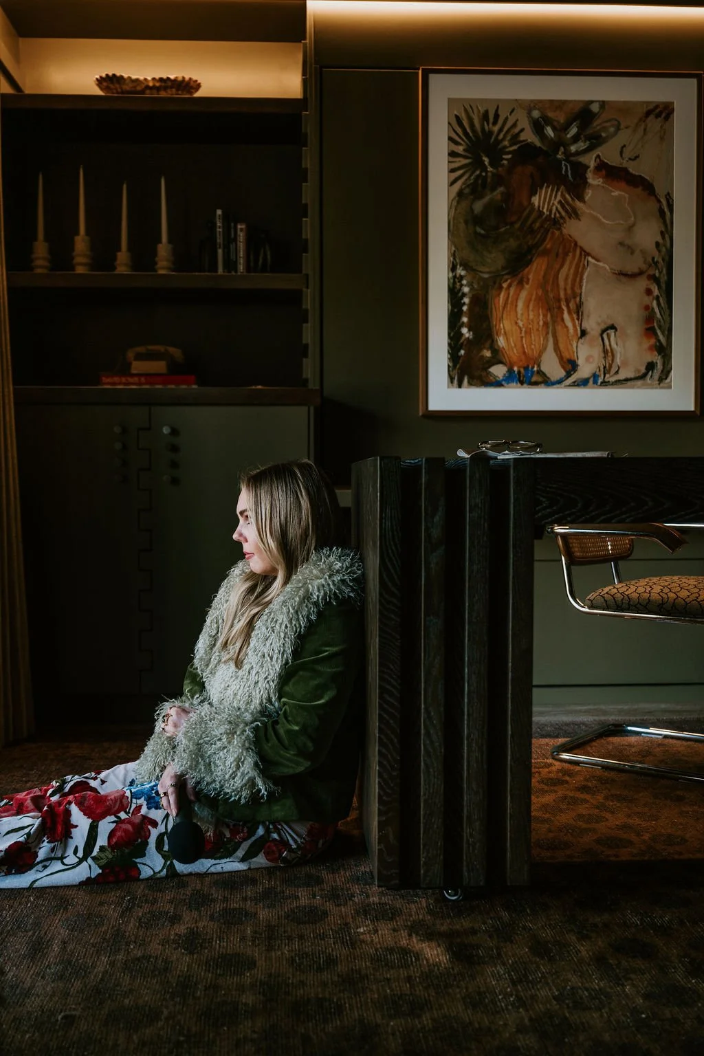 A woman with long blonde hair sitting on the floor beside a dark wooden partition in a room with modern decor, including a large abstract painting on the wall and shelves with books and candles.