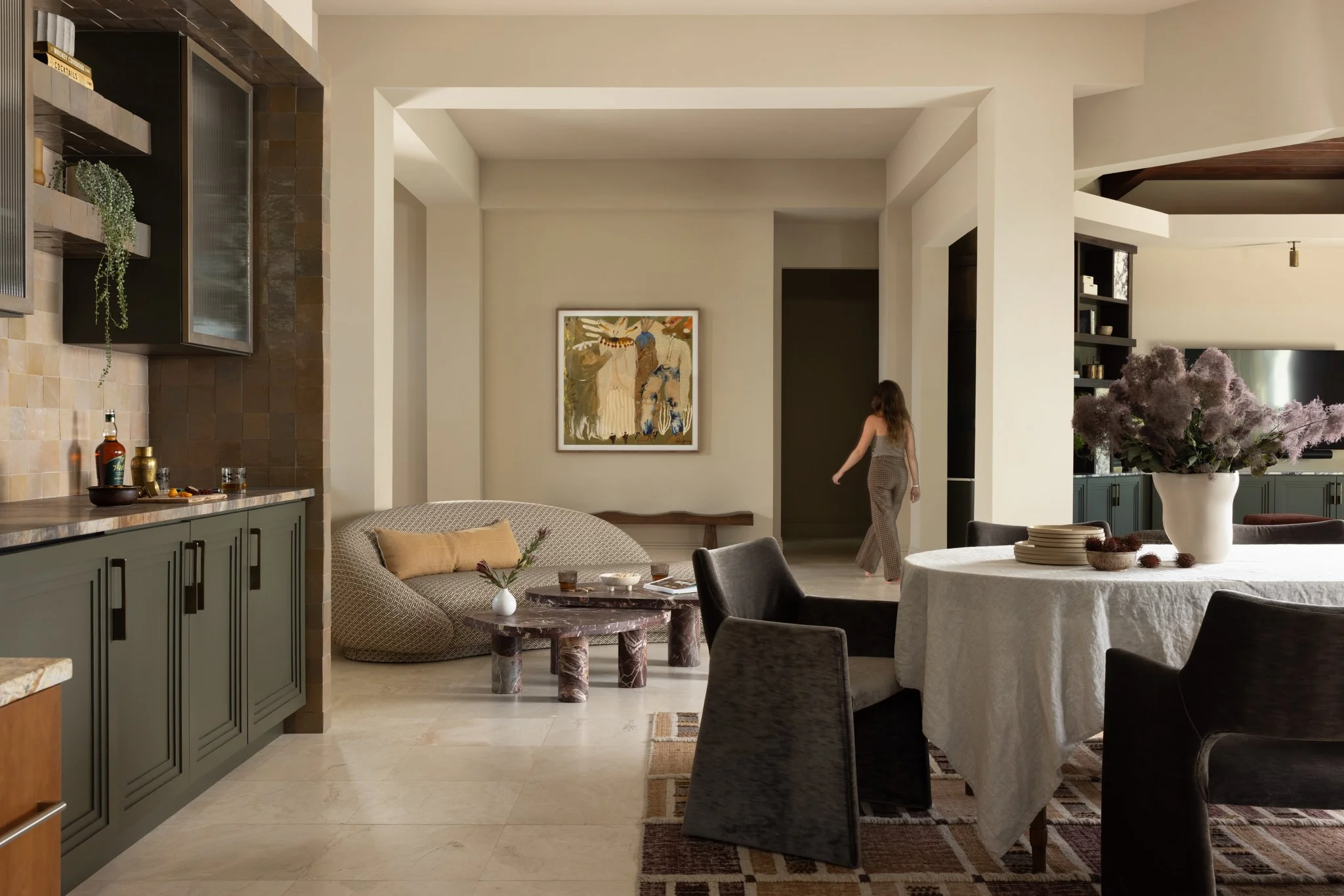 Living room with green custom cabinets, round dining table with white tablecloth and brown chairs, purple flower arrangement in white vase, art on the wall, woman walking, sofa with yellow pillow, coffee table, rug, and cabinetry.