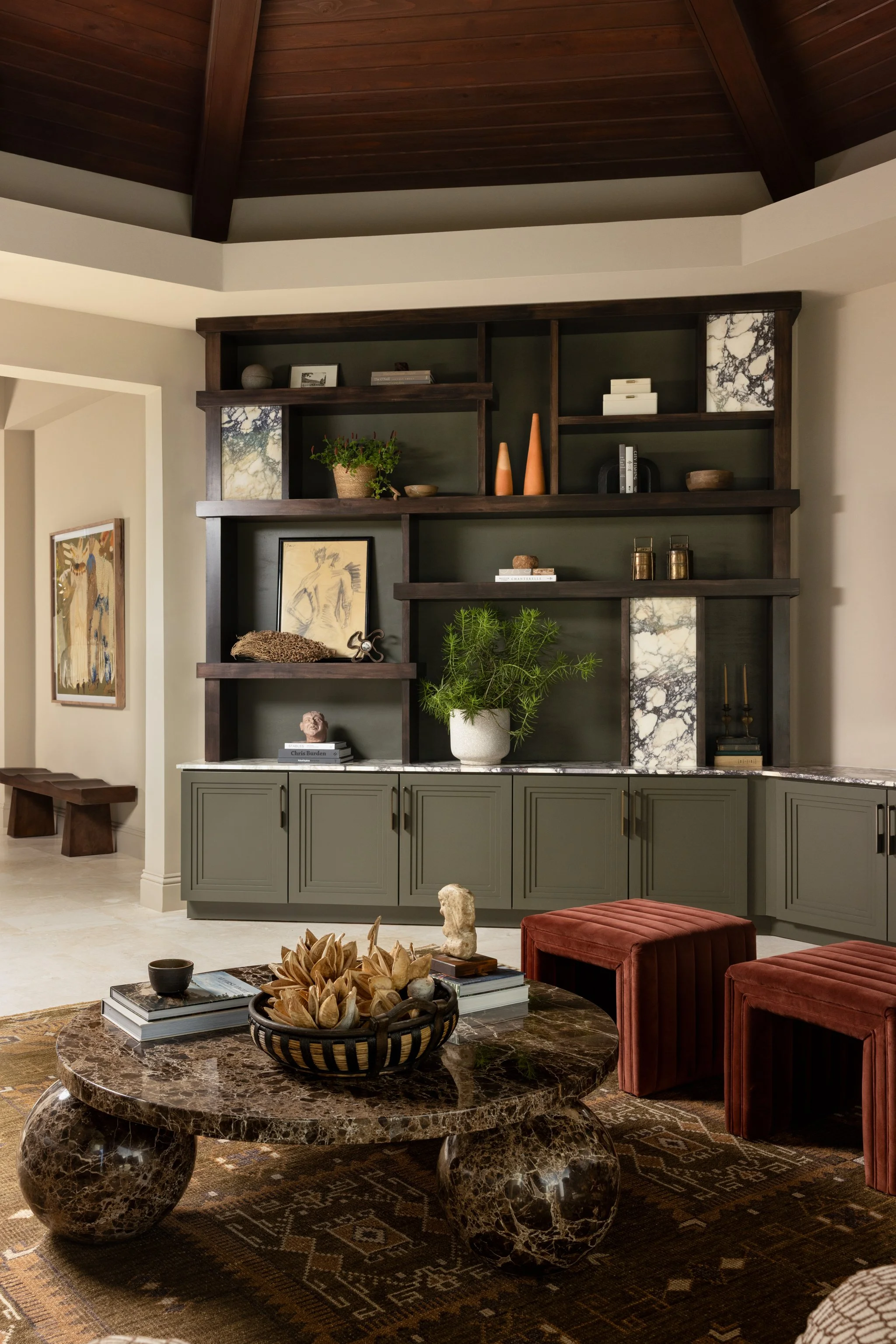 Living room with a built-in dark wood and marble shelving unit, green lower cabinets, a round marble coffee table, and red velvet ottomans, decorated with plants, books, and decorative objects.