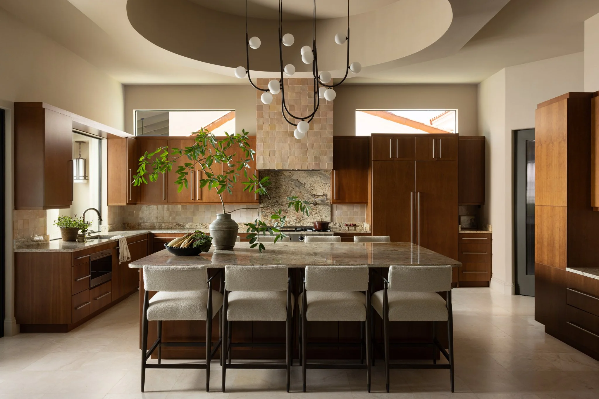 Modern kitchen with wooden cabinets, a large granite island, and a contemporary chandelier above. There are two windows behind the cabinets and a potted plant on the island.