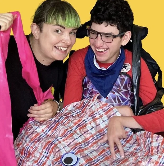 Vicki, a white woman with hair tied back and a green fringe giggles at the camera, she is next to Jonah a white younger man with cool glasses and brown curly hair who is looking at Vicki in a fit of laughter. One of BLINK’s most popular dance props;