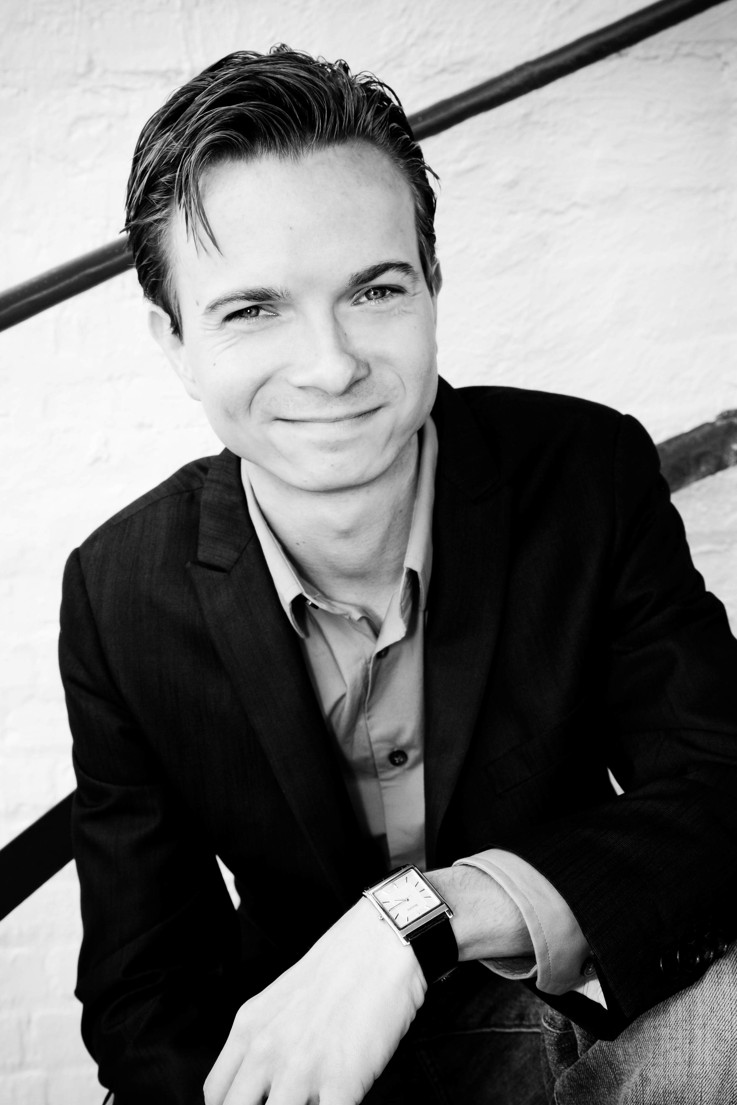 A black and white photo of Wink, a white man, clean shaven with hair parted to one side, not wearing glasses smiling at the camera while sitting in a stairwell and waring a suit, button up shirt, with no tie